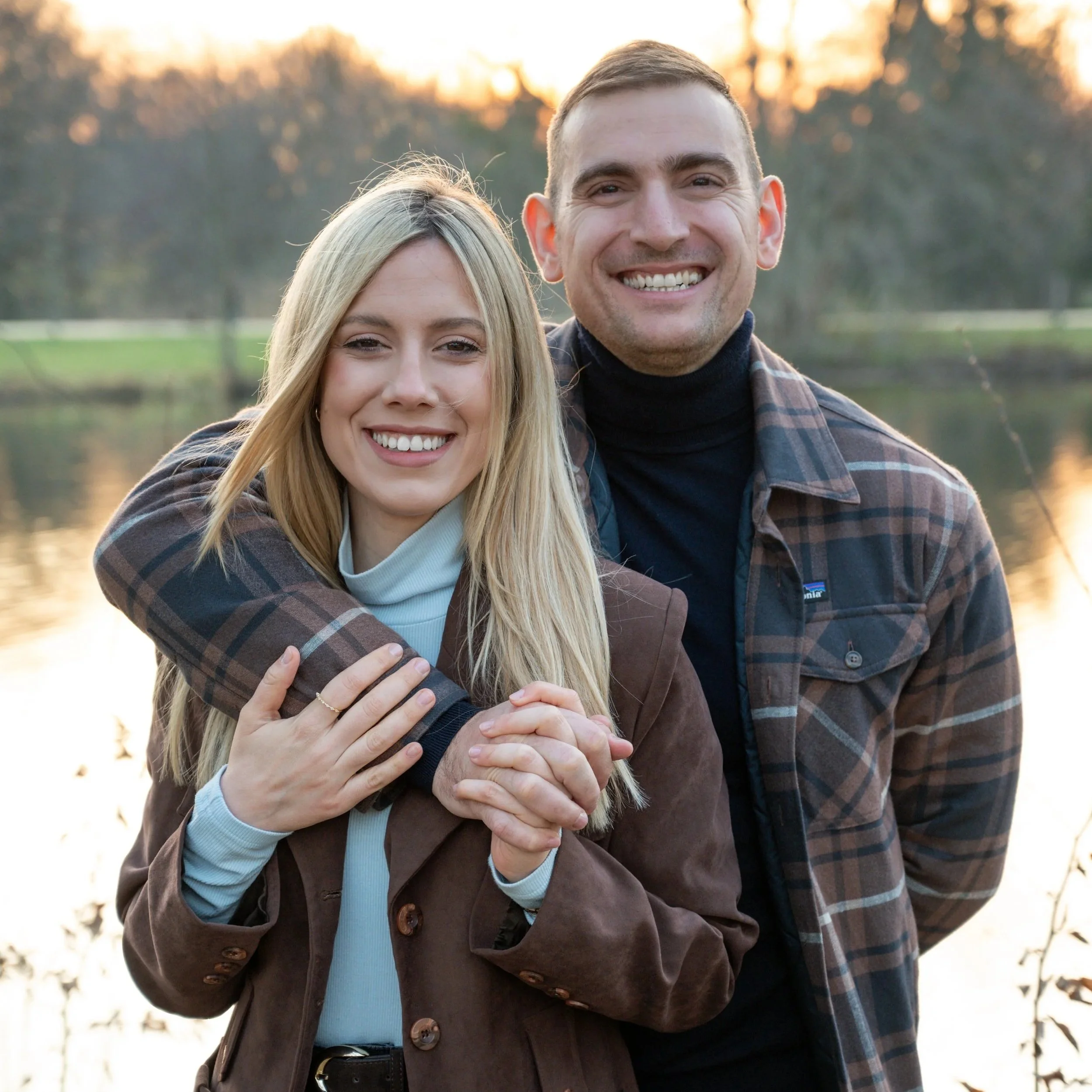 A smiling couple stands outdoors near a body of water during sunset, with the man hugging the woman from behind.