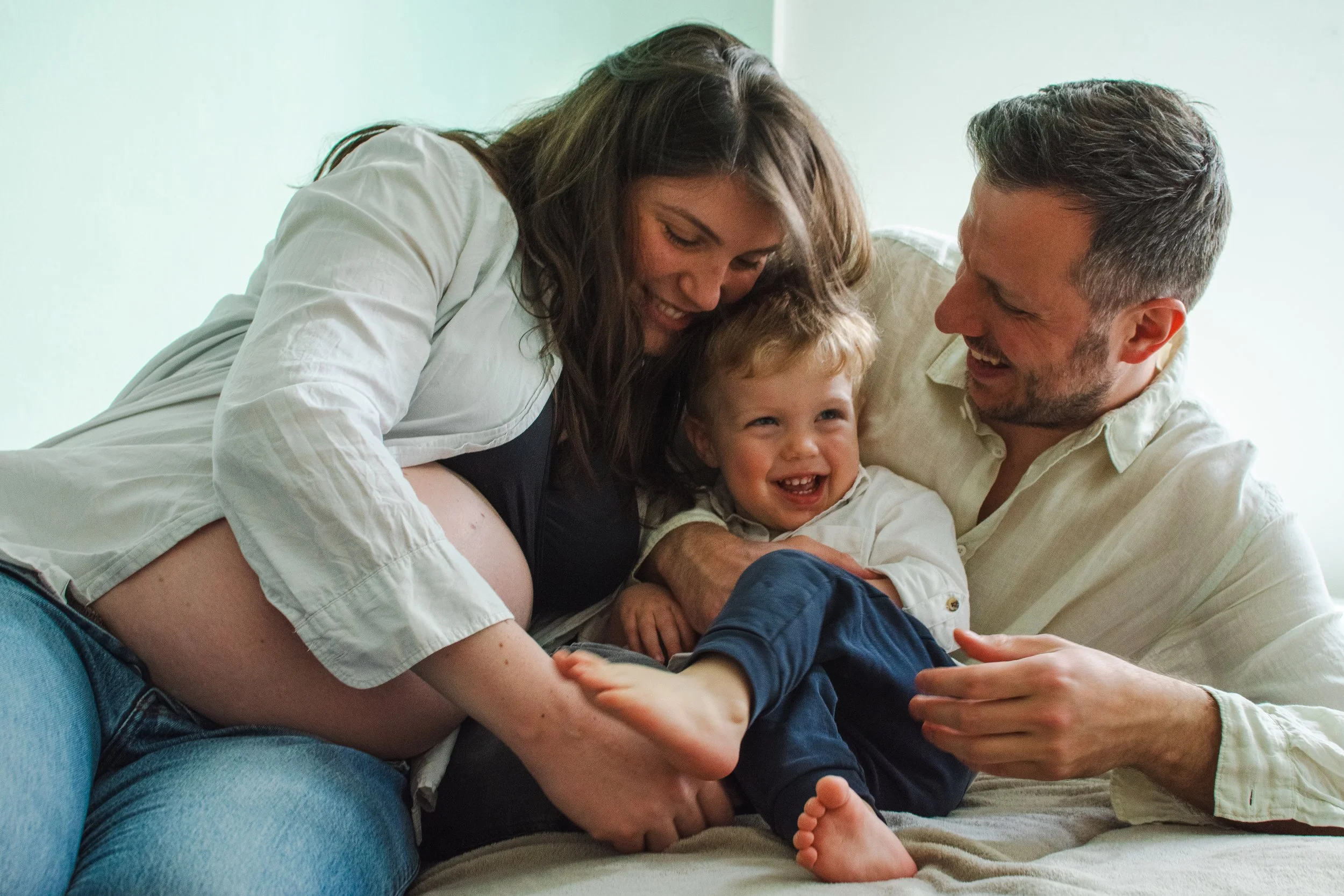 A happy family of three, including a pregnant woman, is playing and laughing together on a sofa in a cozy living room.
