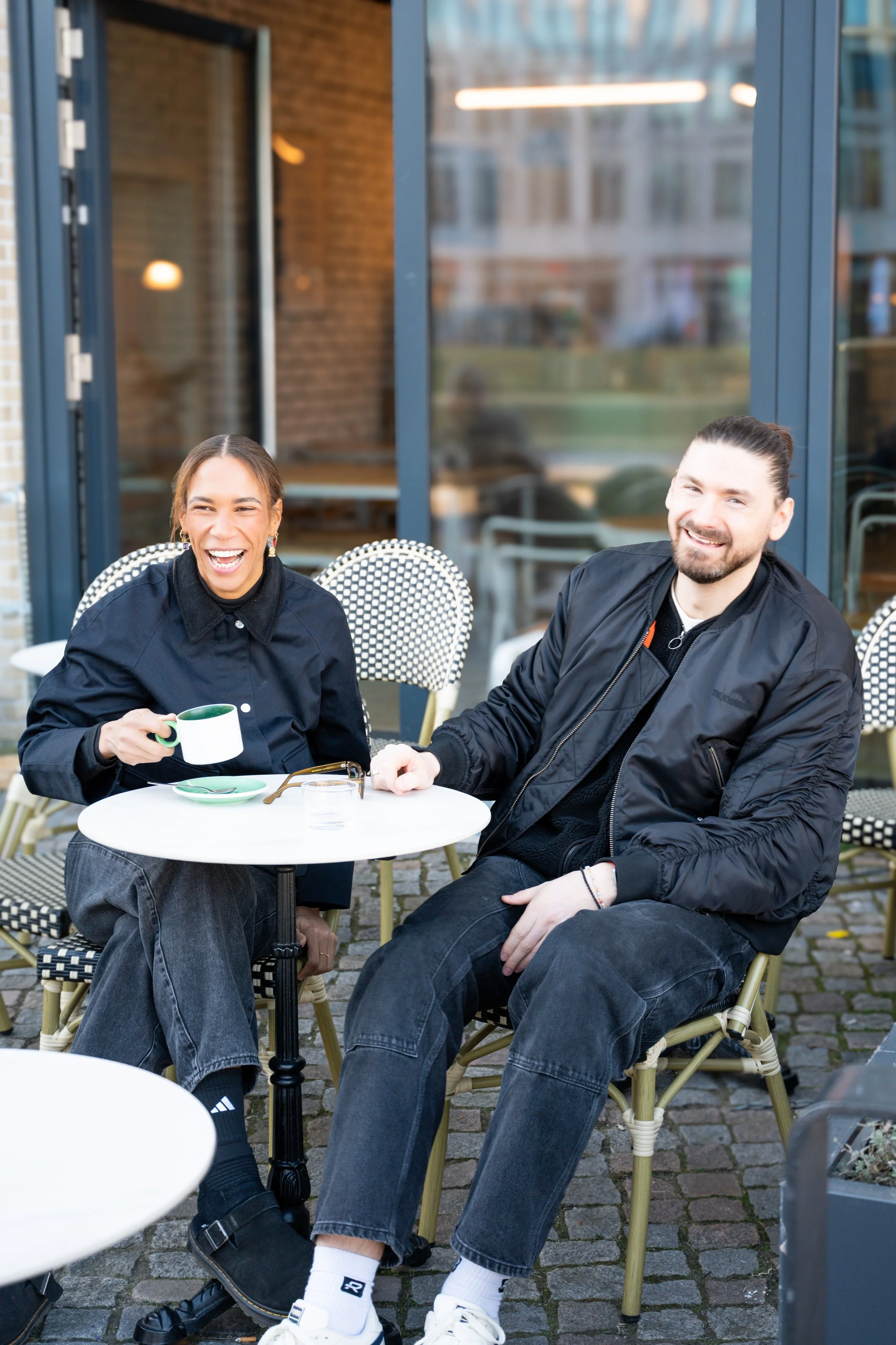 Two people sitting at an outdoor cafe table, laughing and enjoying coffee.