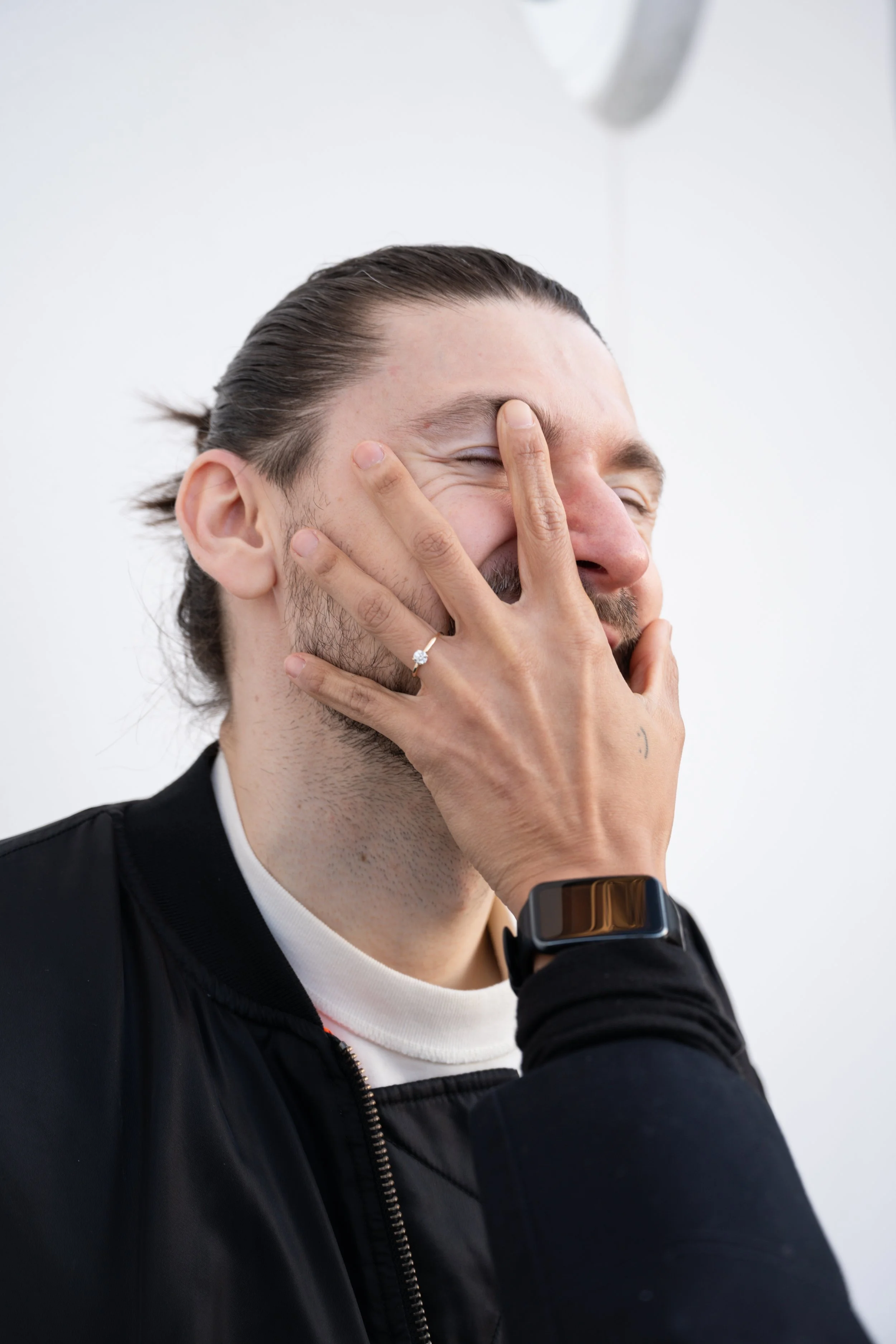 A man with long hair tied back, wearing a black jacket and a smartwatch, laughing while covering his face with his hand.