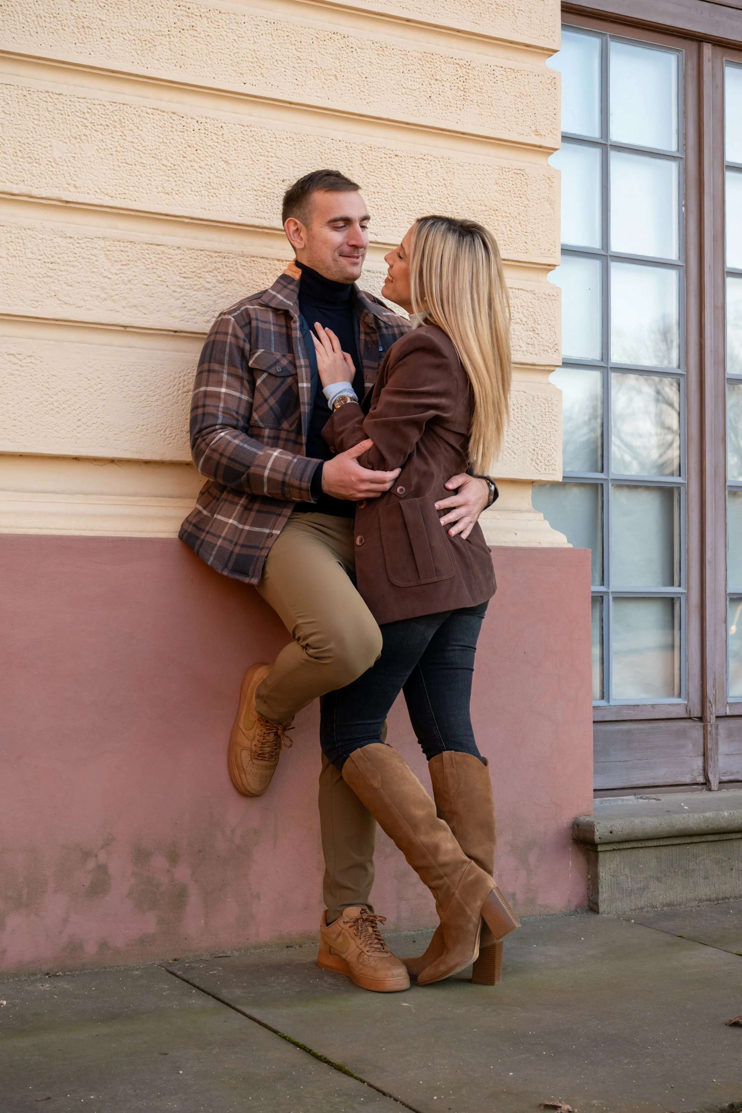 A couple standing close together outdoors near a beige building with large windows, embracing each other and smiling.