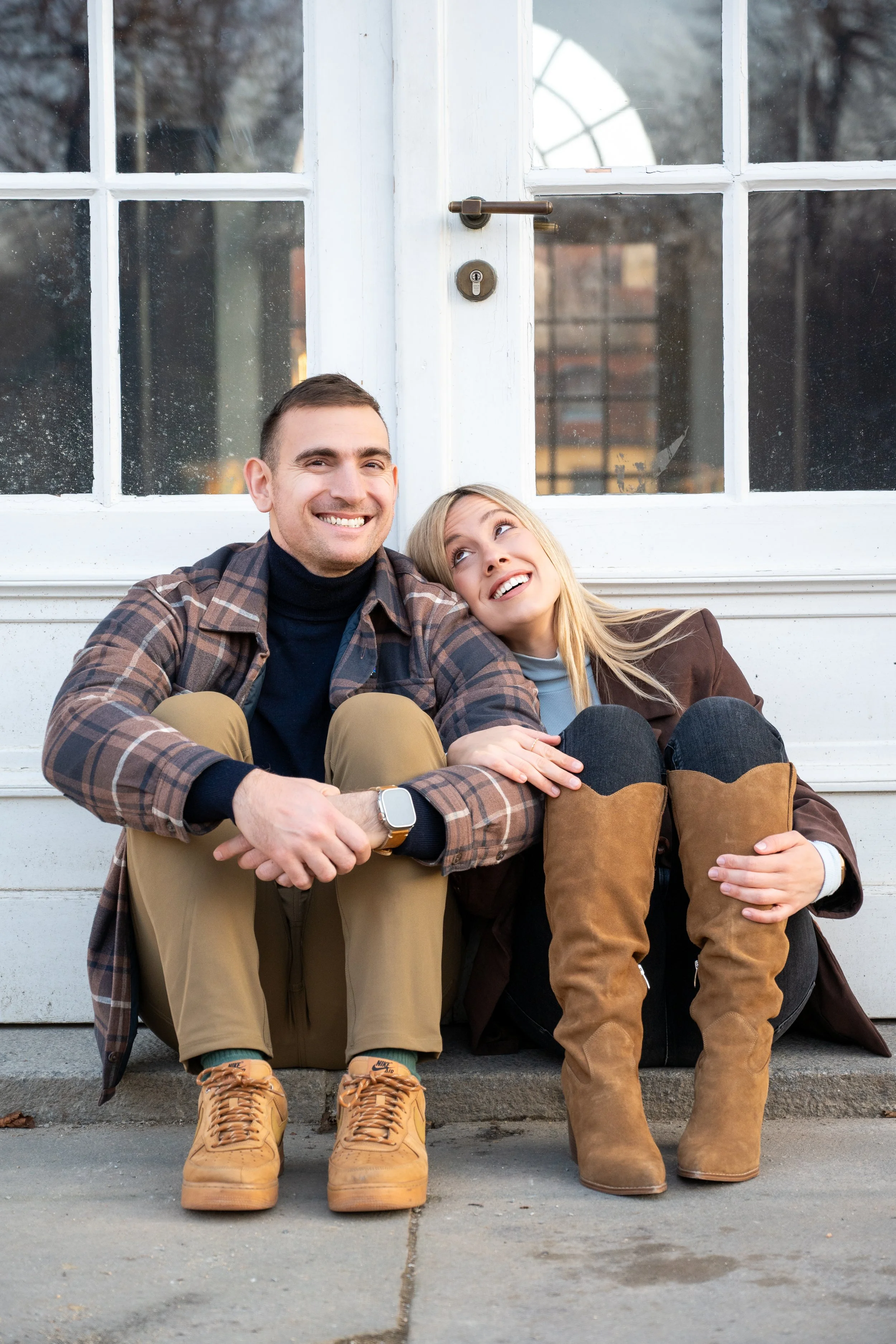 A smiling young man and woman sitting close together on a sidewalk in front of a white building with a glass door. The man is wearing a plaid shirt, black turtleneck, and tan boots, while the woman is wearing a brown jacket, black pants, and tan boot