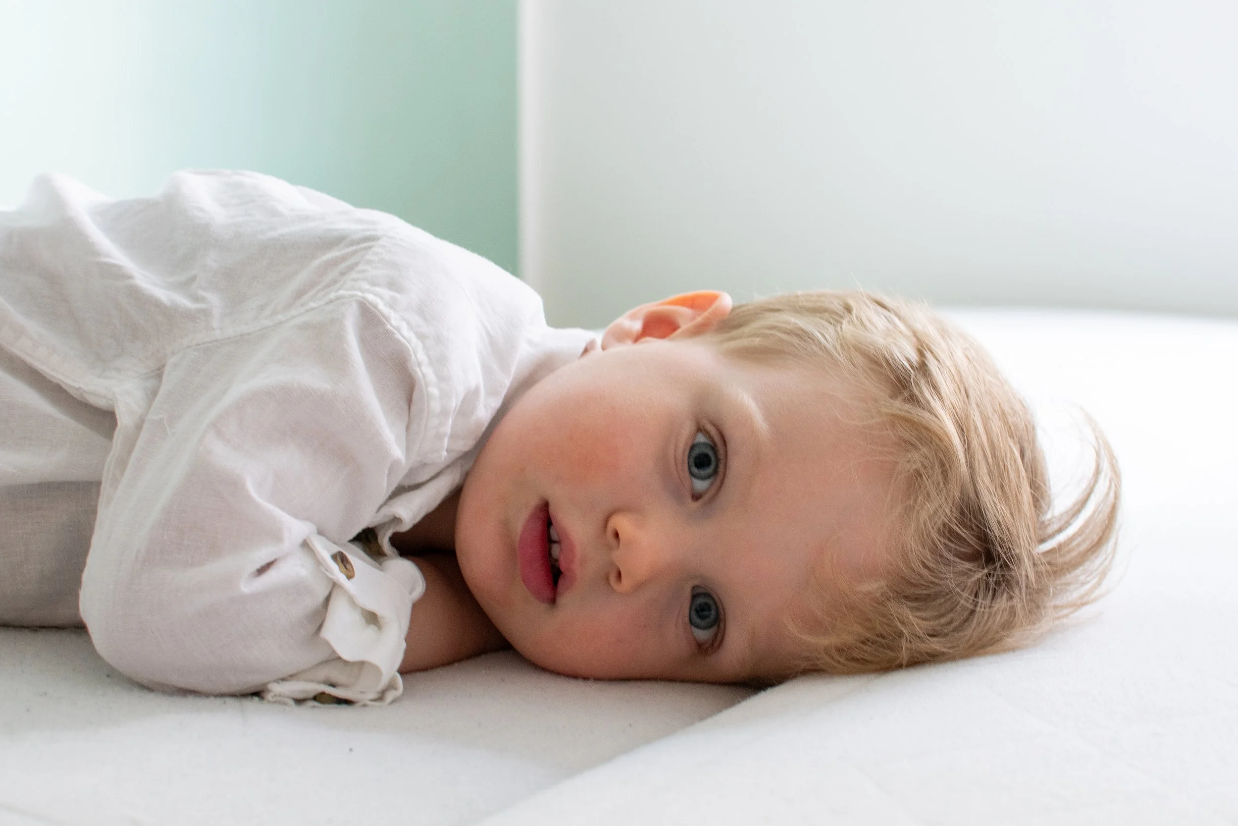 Young boy with blonde hair lying on a white bed, looking toward the camera with a curious expression.
