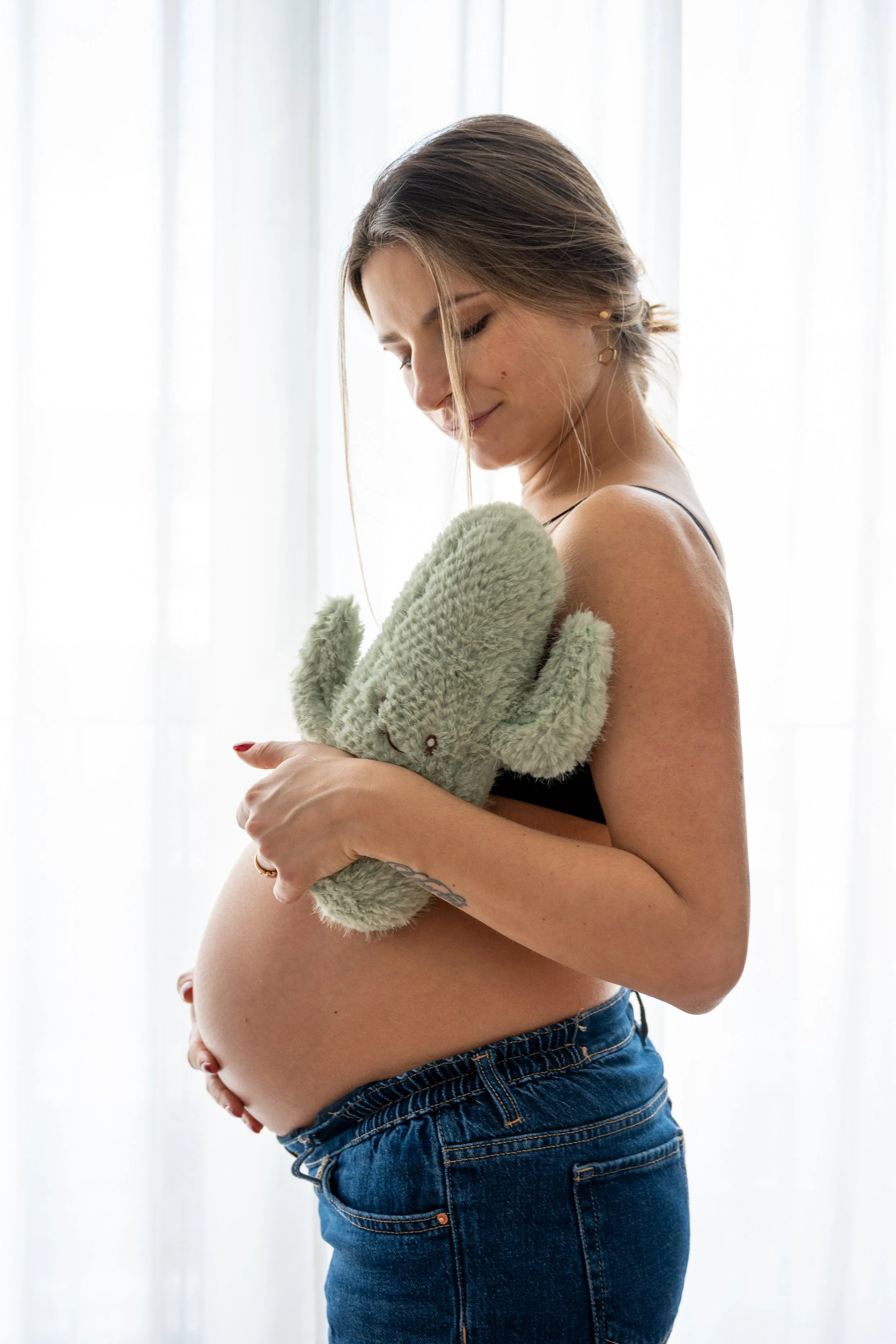 A pregnant woman with light brown hair, wearing a black top and blue jeans, holding a green plush dinosaur toy close to her chest and looking down at her belly with a gentle expression, standing in front of a sheer white curtain.