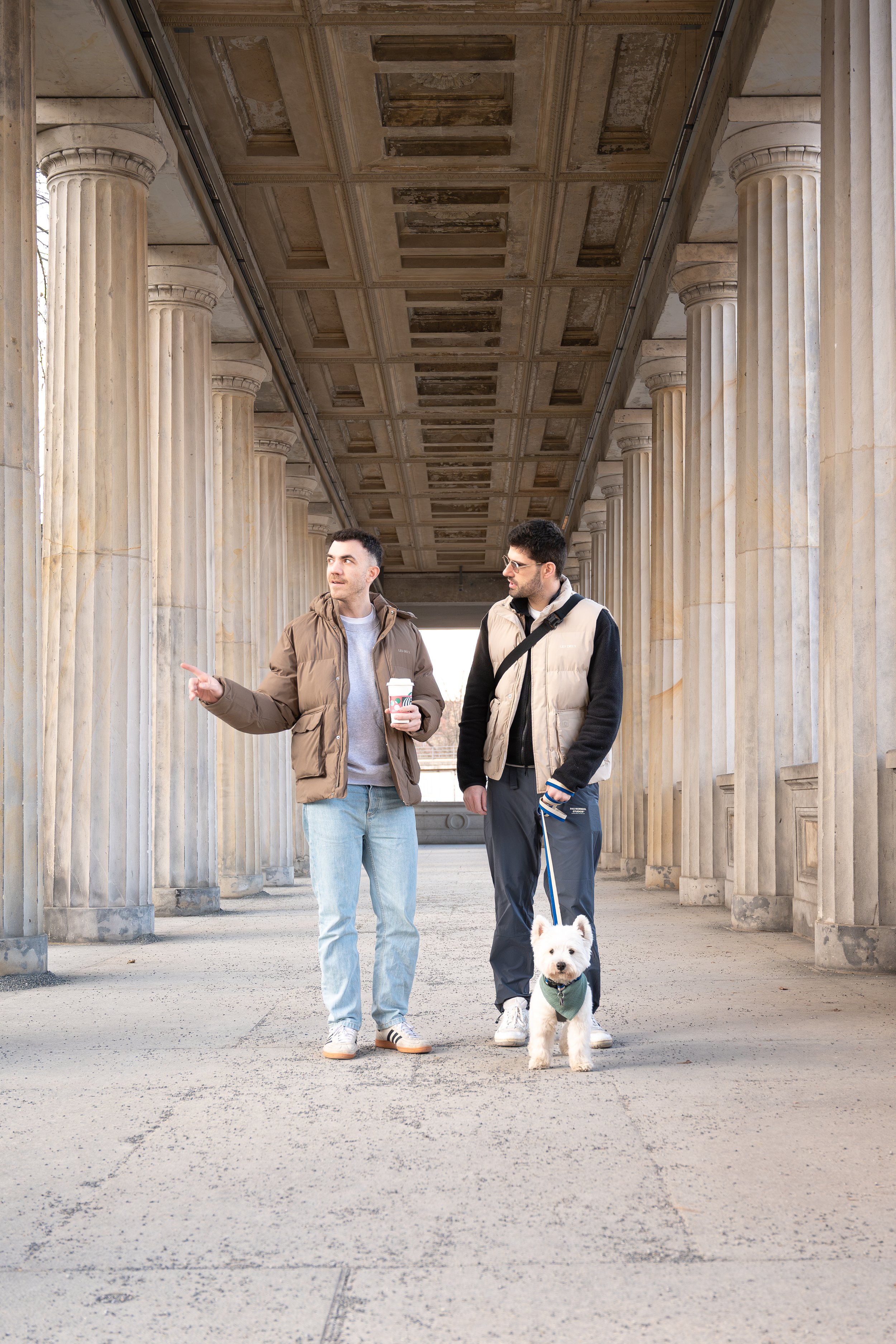 Two men walking and talking under a colonnade, one holding a coffee cup and the other with a dog on a leash.