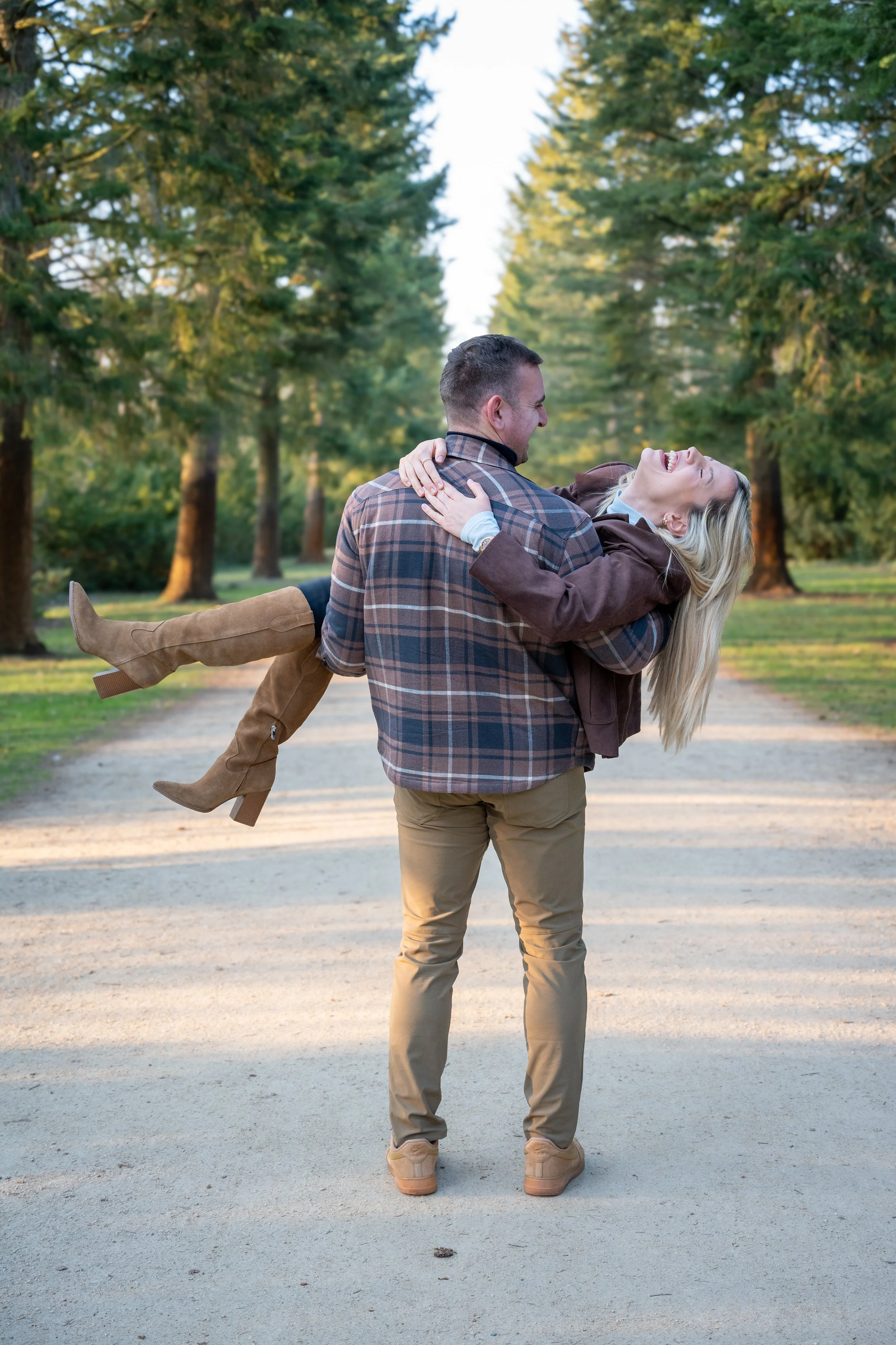 A man is holding a woman in his arms in a park with trees, both women are smiling and laughing.