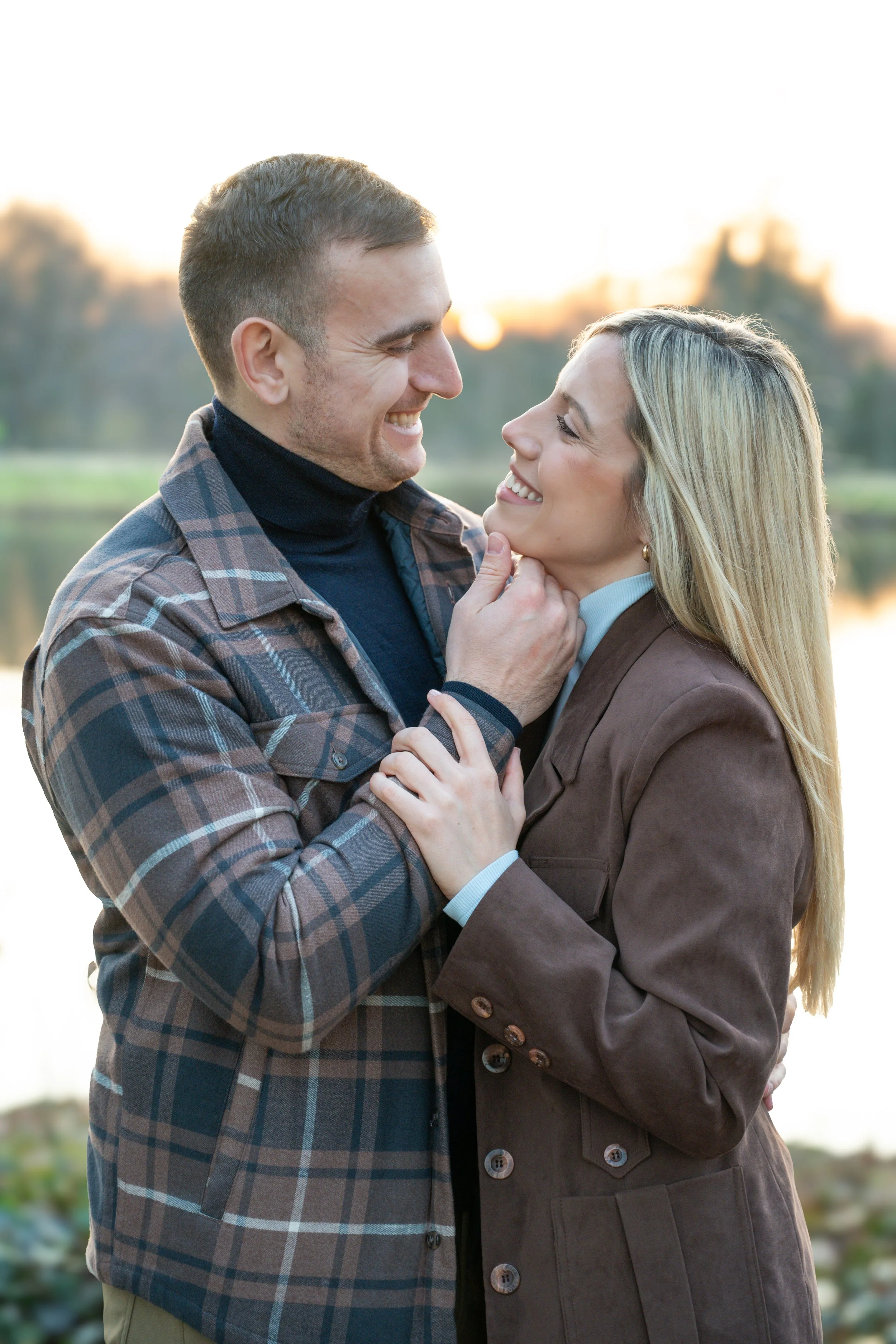 A smiling man and woman embracing outdoors during sunset, with a body of water and trees in the background.