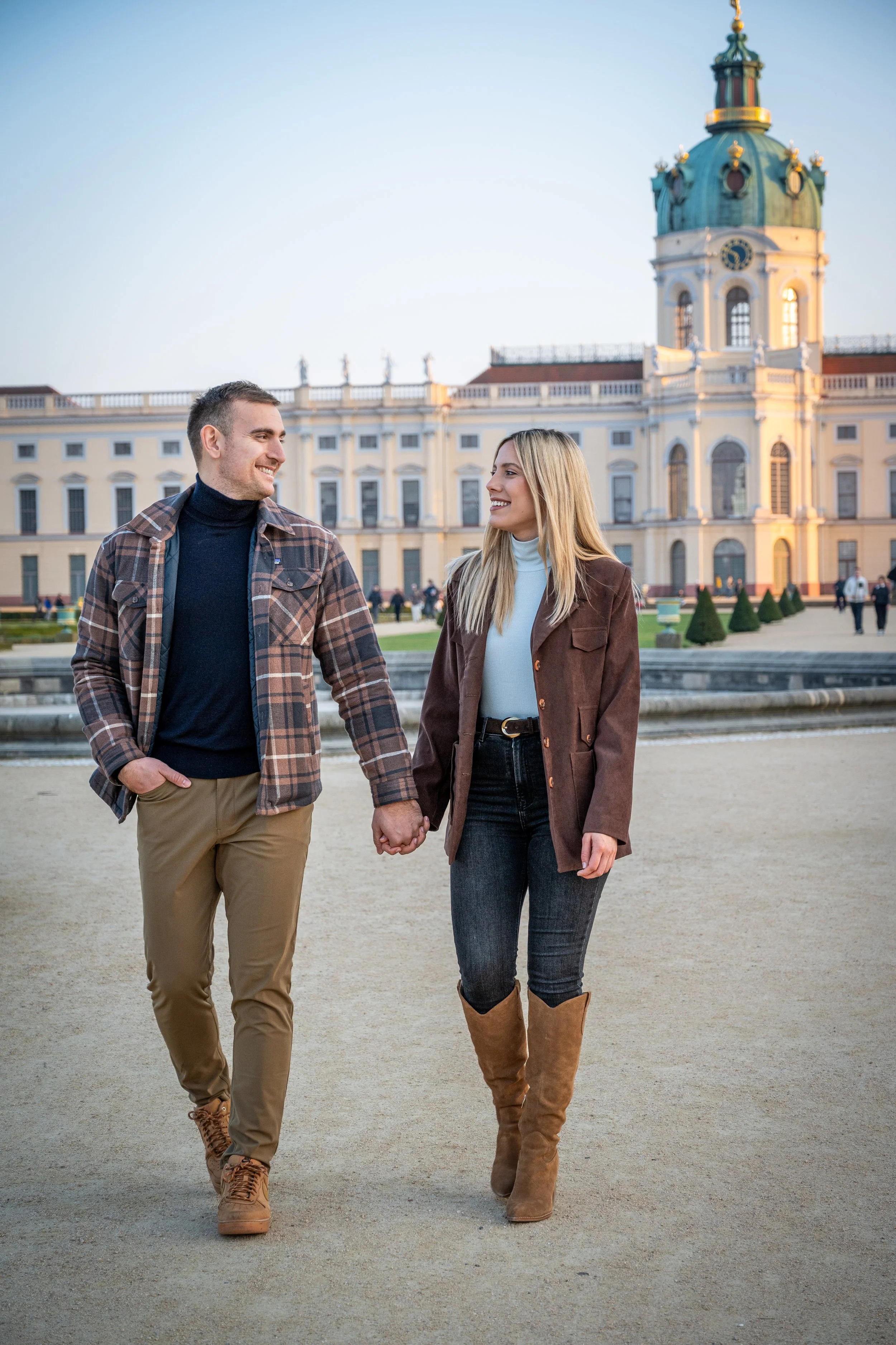 A young couple walking hand-in-hand in front of a historic building with a green-domed clock tower, smiling at each other.