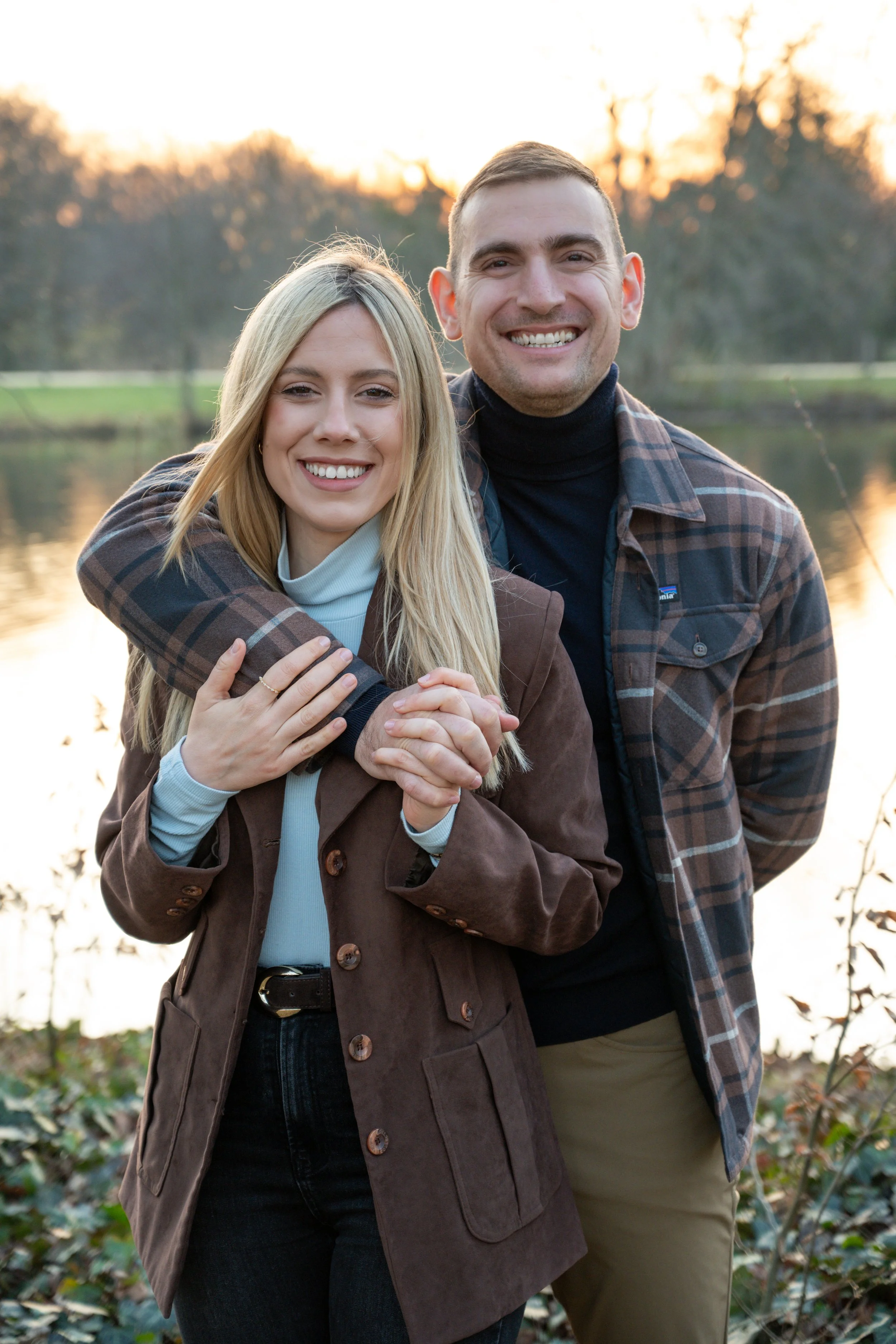 A smiling couple outdoors near a lake during sunset, with trees in the background, embracing and holding hands.
