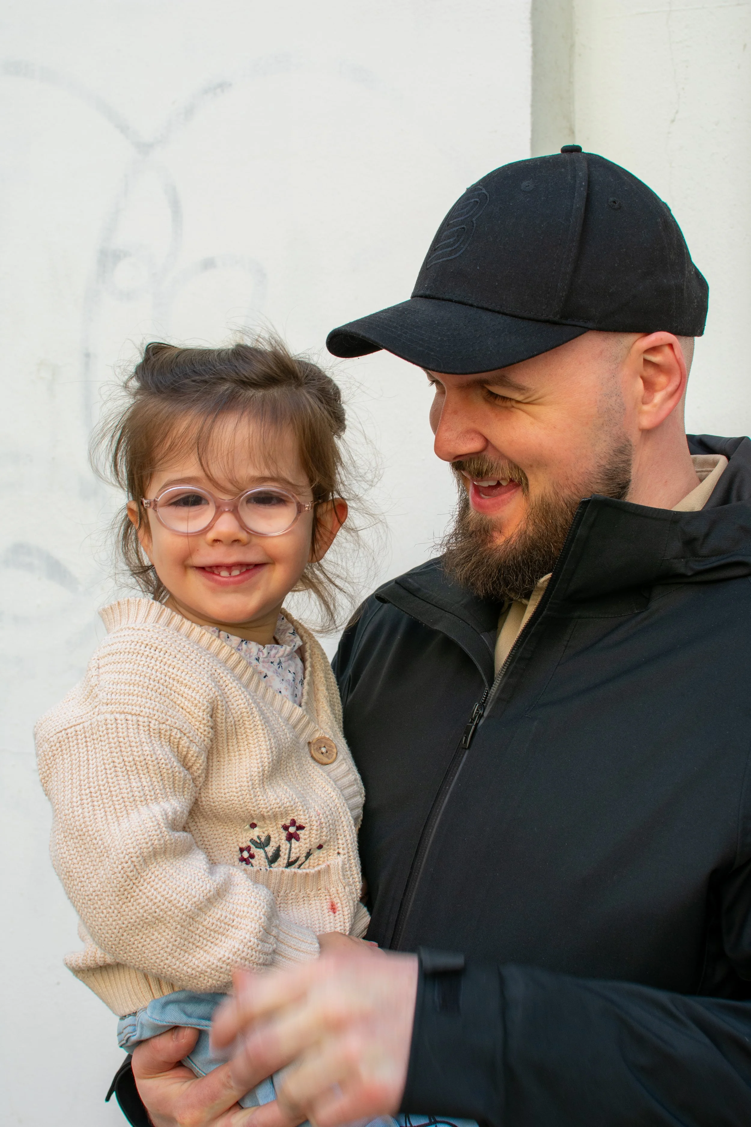 A man and a young girl are smiling and holding hands, standing close together against a plain white wall.