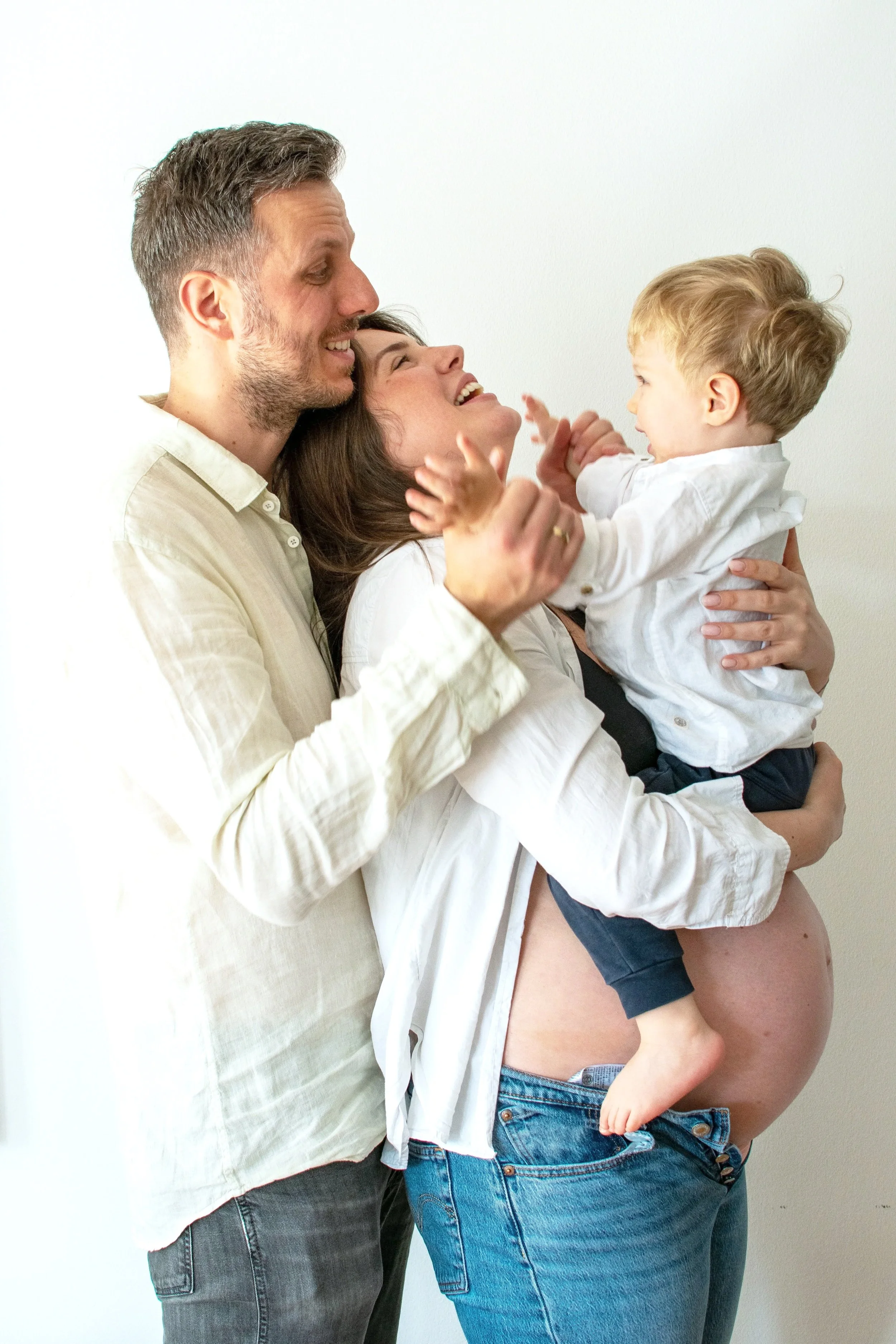 Family of three celebrating pregnancy, with father, mother, and young son, smiling and embracing against a plain white background.