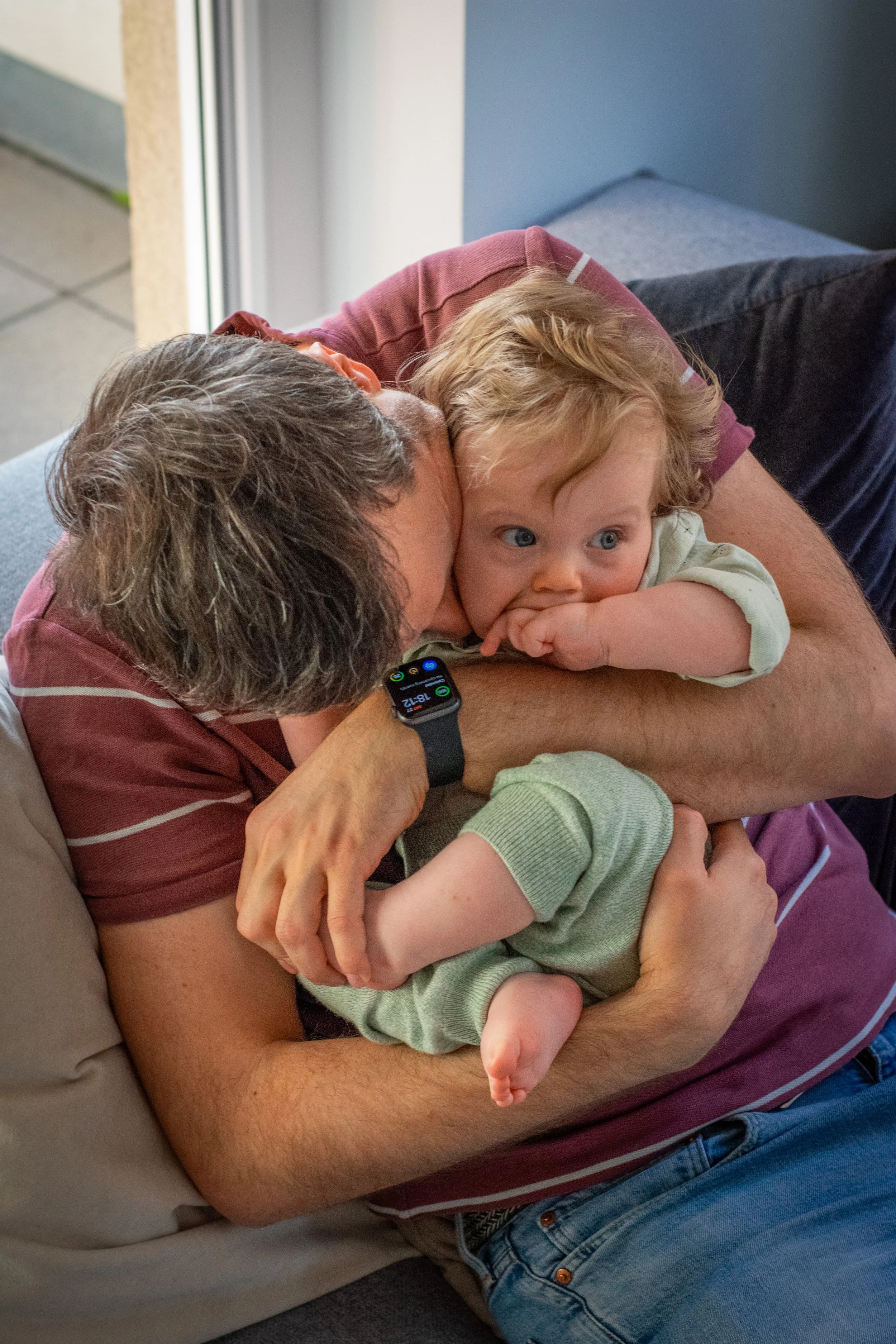 A man with gray hair wearing a striped red shirt is lying on a sofa, holding two young children. One child, with blond hair and wide eyes, is resting their head on the man's arm, while the other, with lighter hair, is being held and has a finger in t
