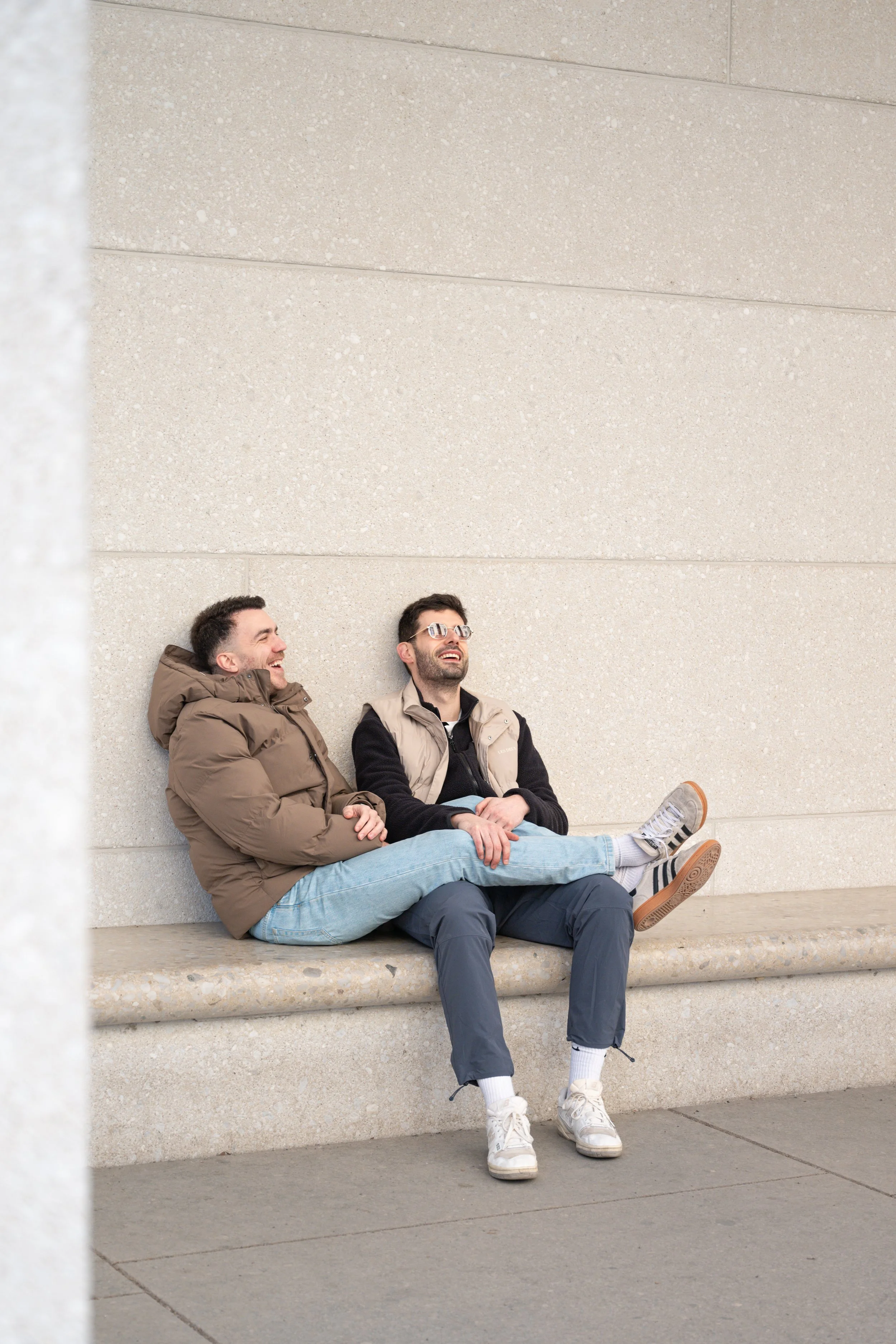 Two young men sitting on a bench against a concrete wall, one with a brown jacket and the other with a beige vest, both smiling and laughing.