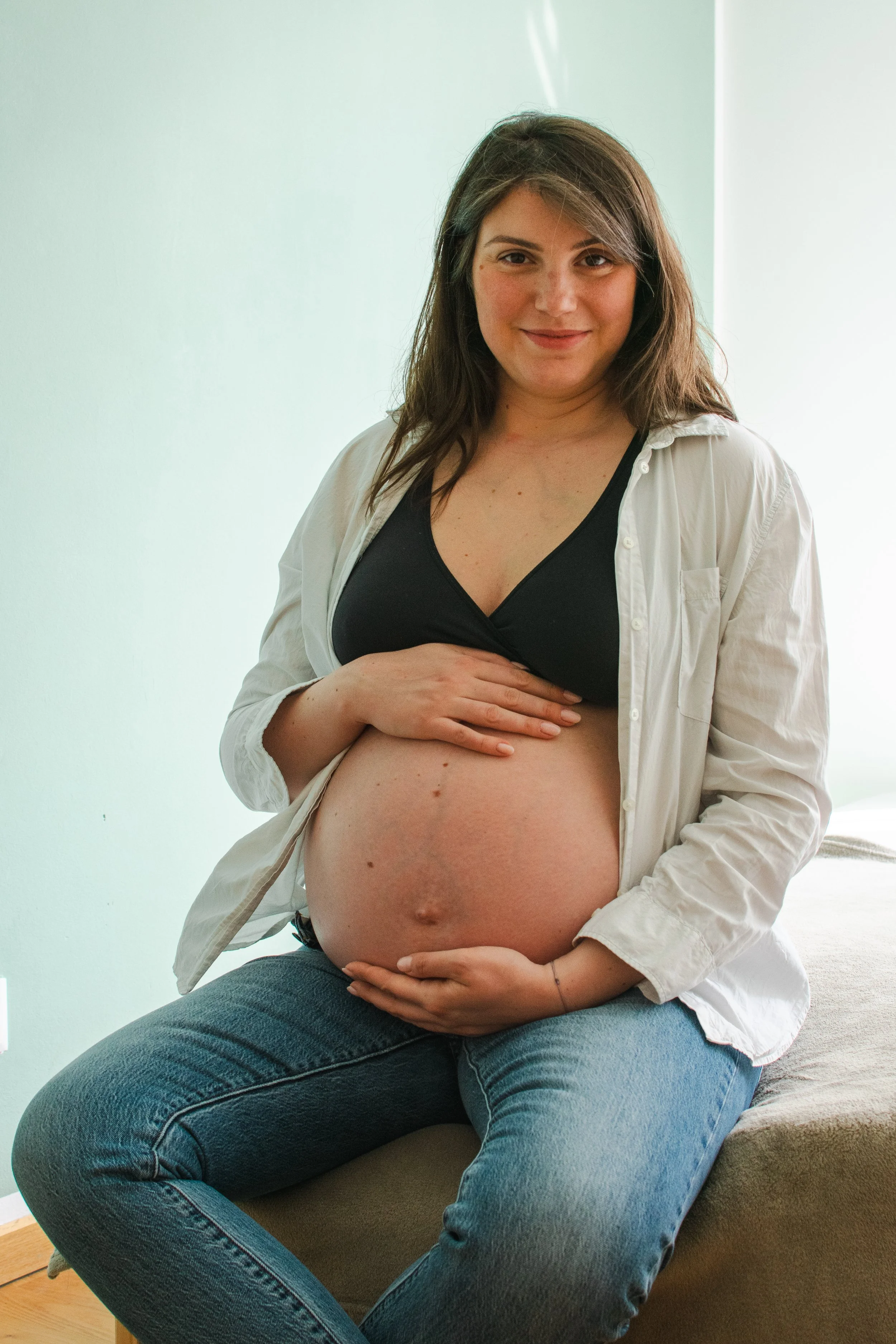 A pregnant woman smiling while sitting on a bed, wearing a black top, an unbuttoned white shirt, and blue jeans, holding her belly with both hands.