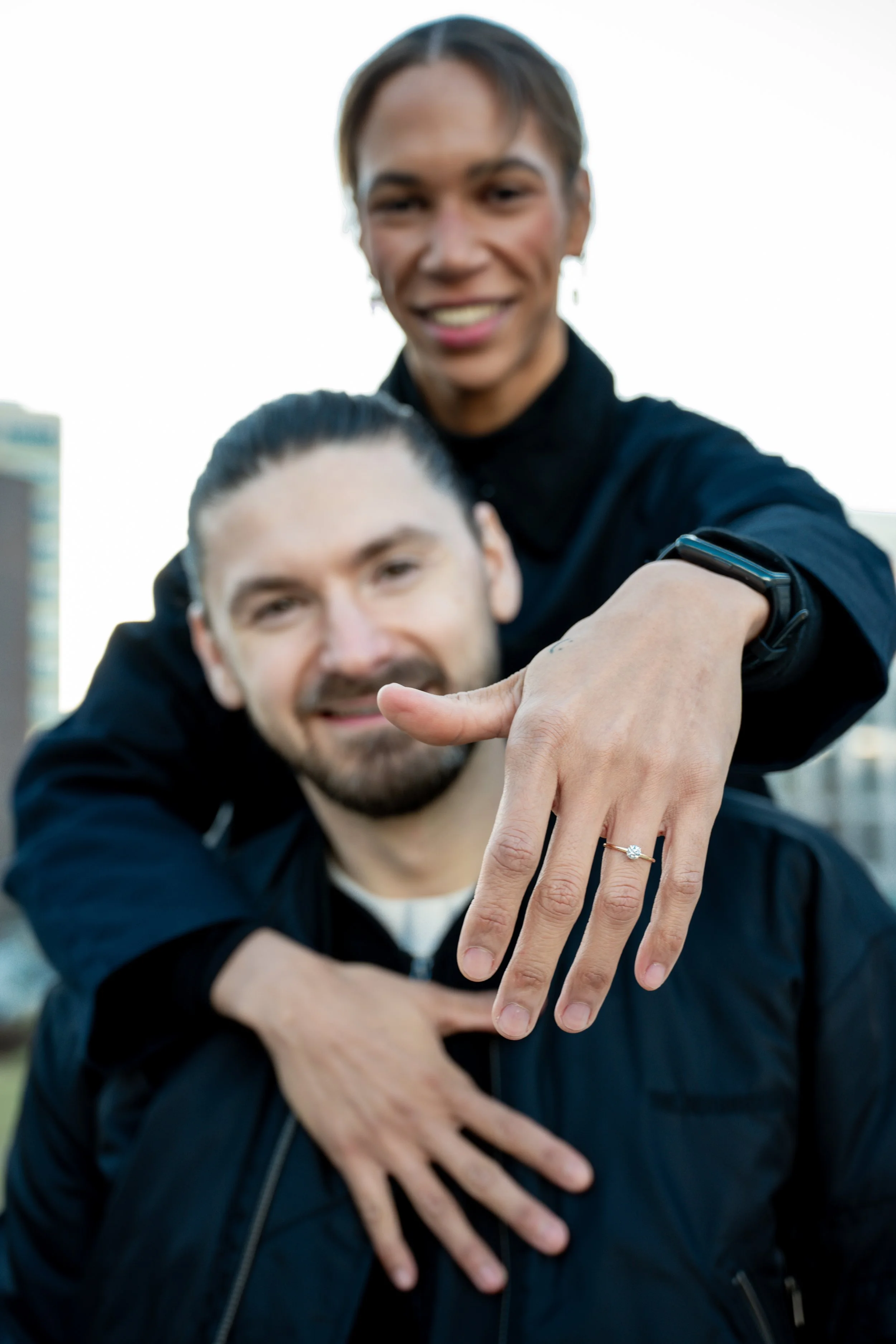 Two men outdoors, one with long hair and the other with short hair, smiling at the camera. The man with long hair is showing a ring on his finger, indicating an engagement or marriage.