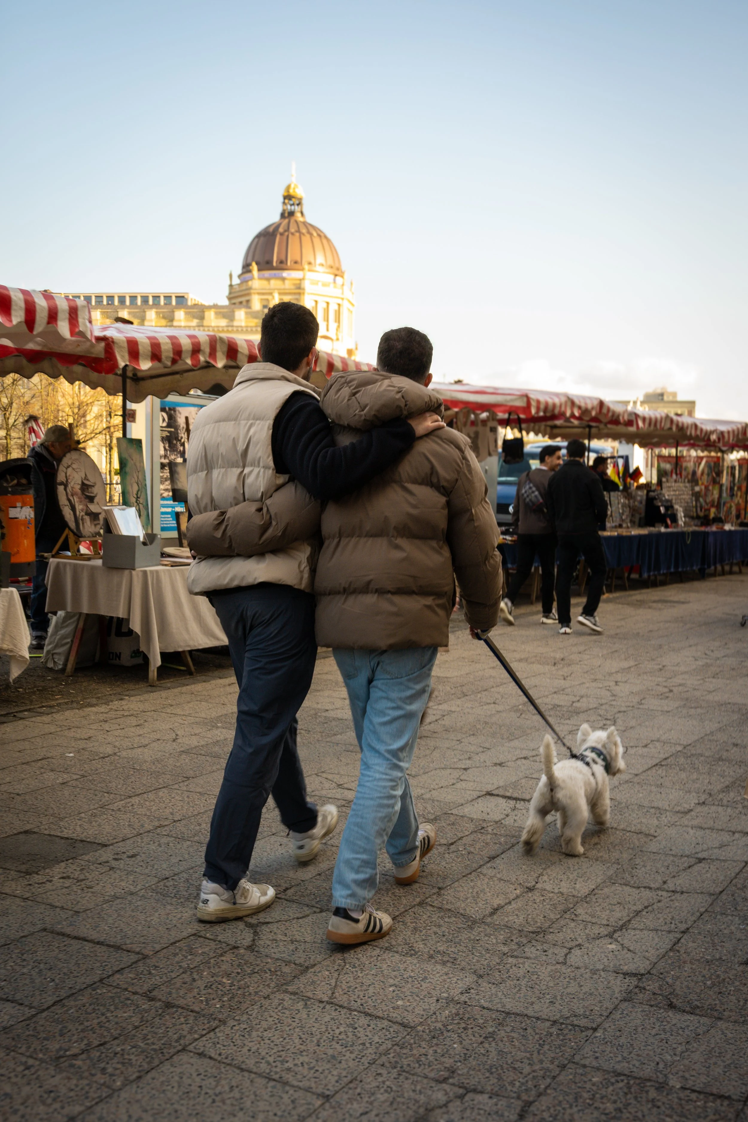 Two men walk through an outdoor market with a small dog on a leash, market stalls with striped red and white awnings, and a historic domed building in the background.