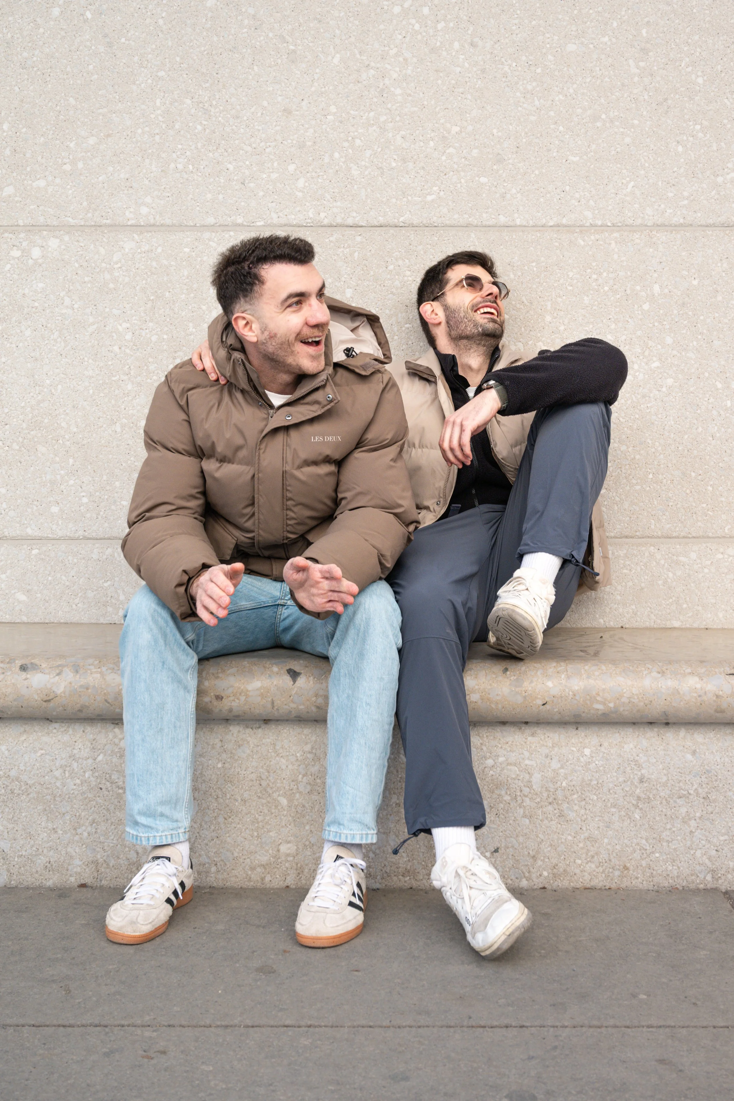 Two young men sitting on a concrete bench against a beige wall, laughing and enjoying each other's company while wearing casual outdoor clothing.
