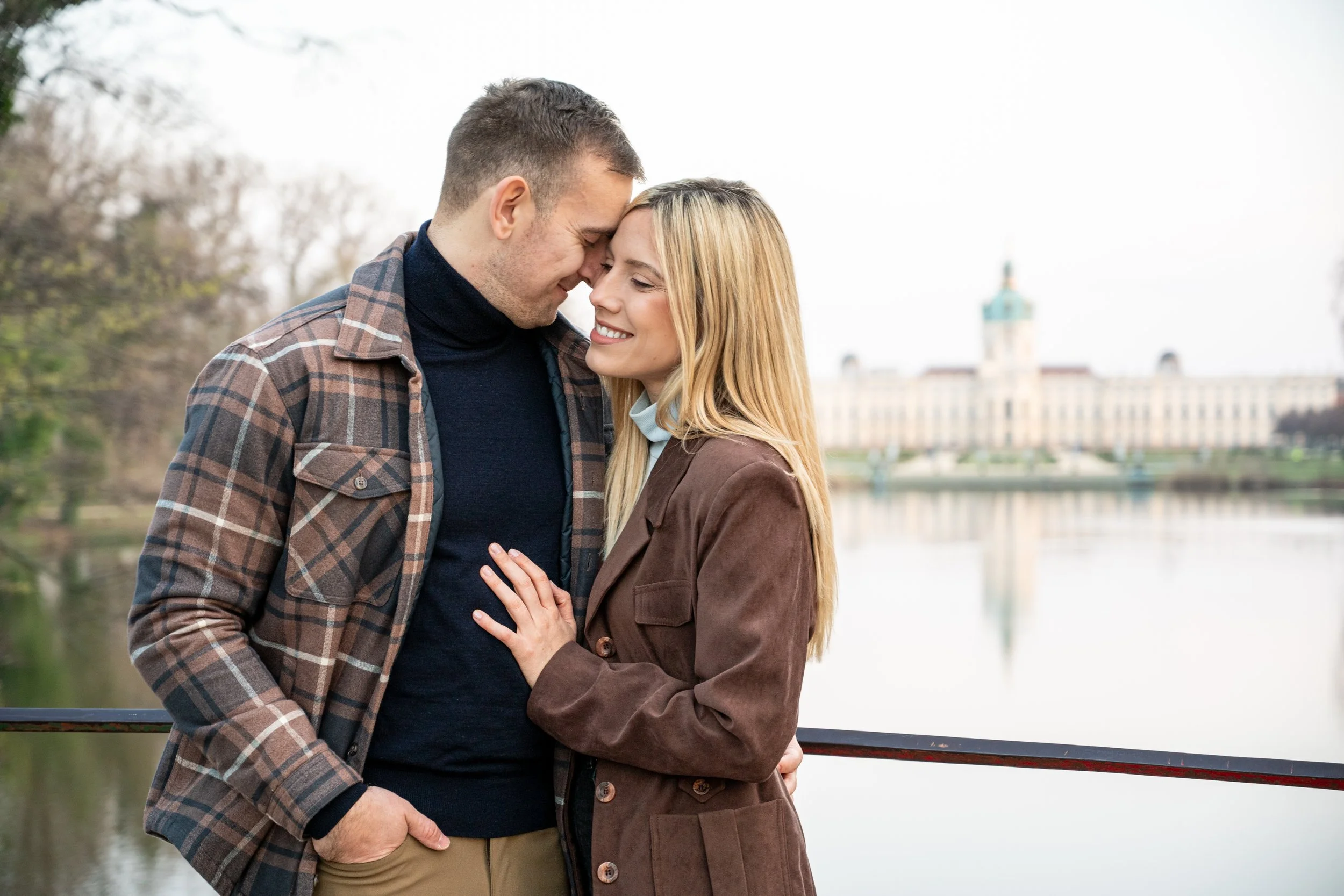 A young couple sharing a tender moment by a river, with historical building in the background.