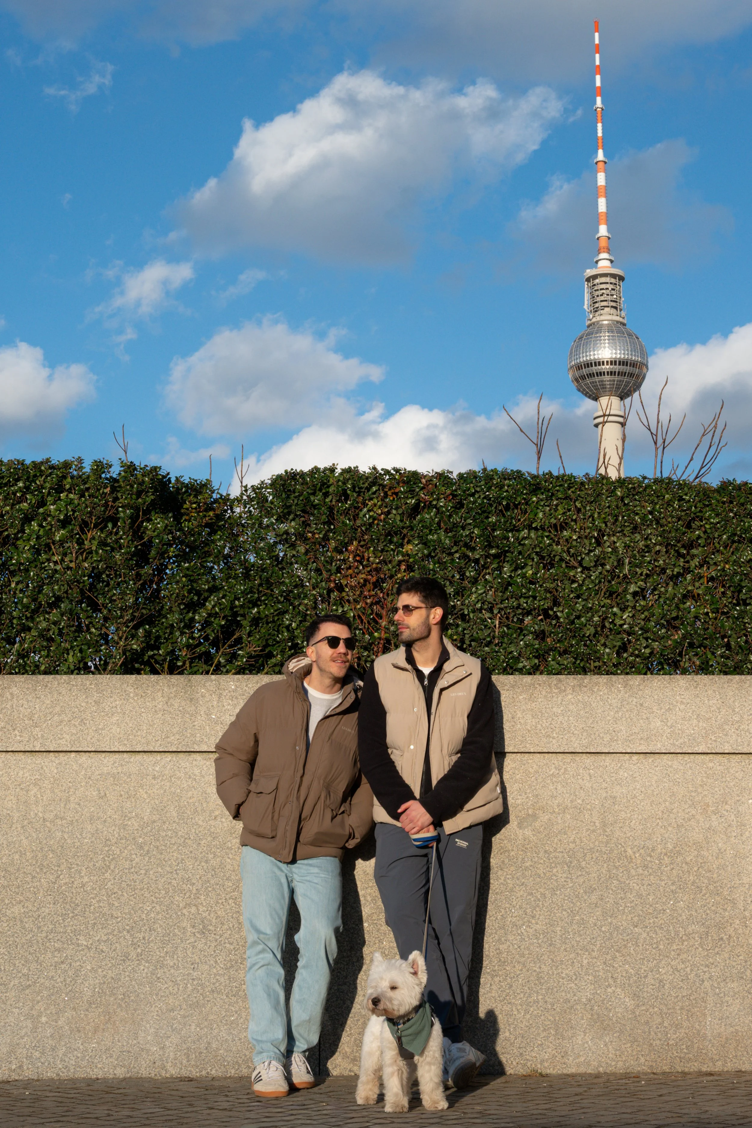 Two men standing outdoors with a white dog on a leash, in front of a hedge and Berlin TV Tower against a blue sky with white clouds.