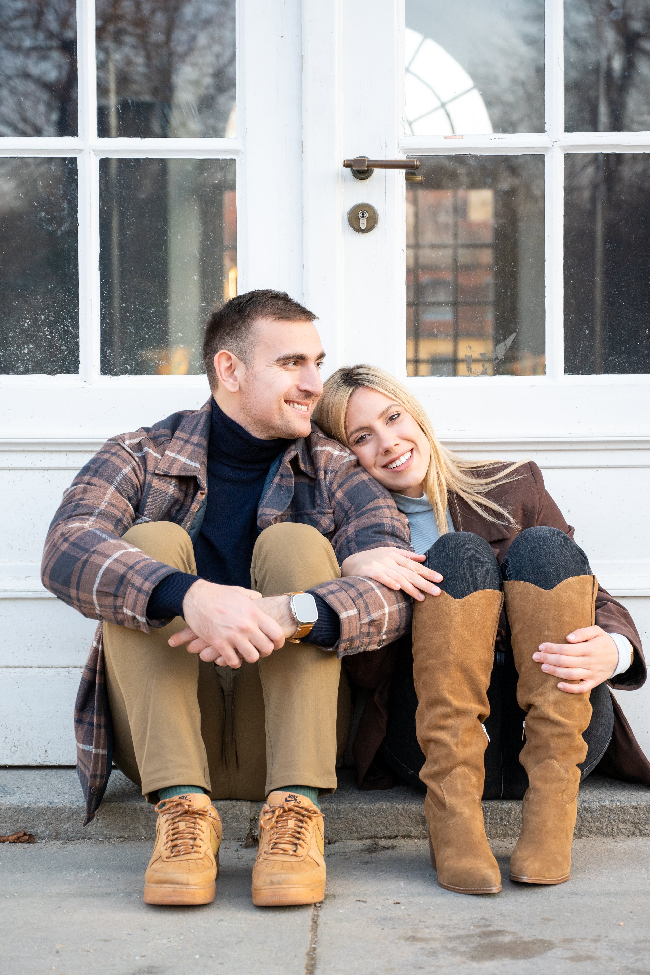 A smiling young man and woman sitting on a step outside a white wooden building, resting against a glass-paneled door.