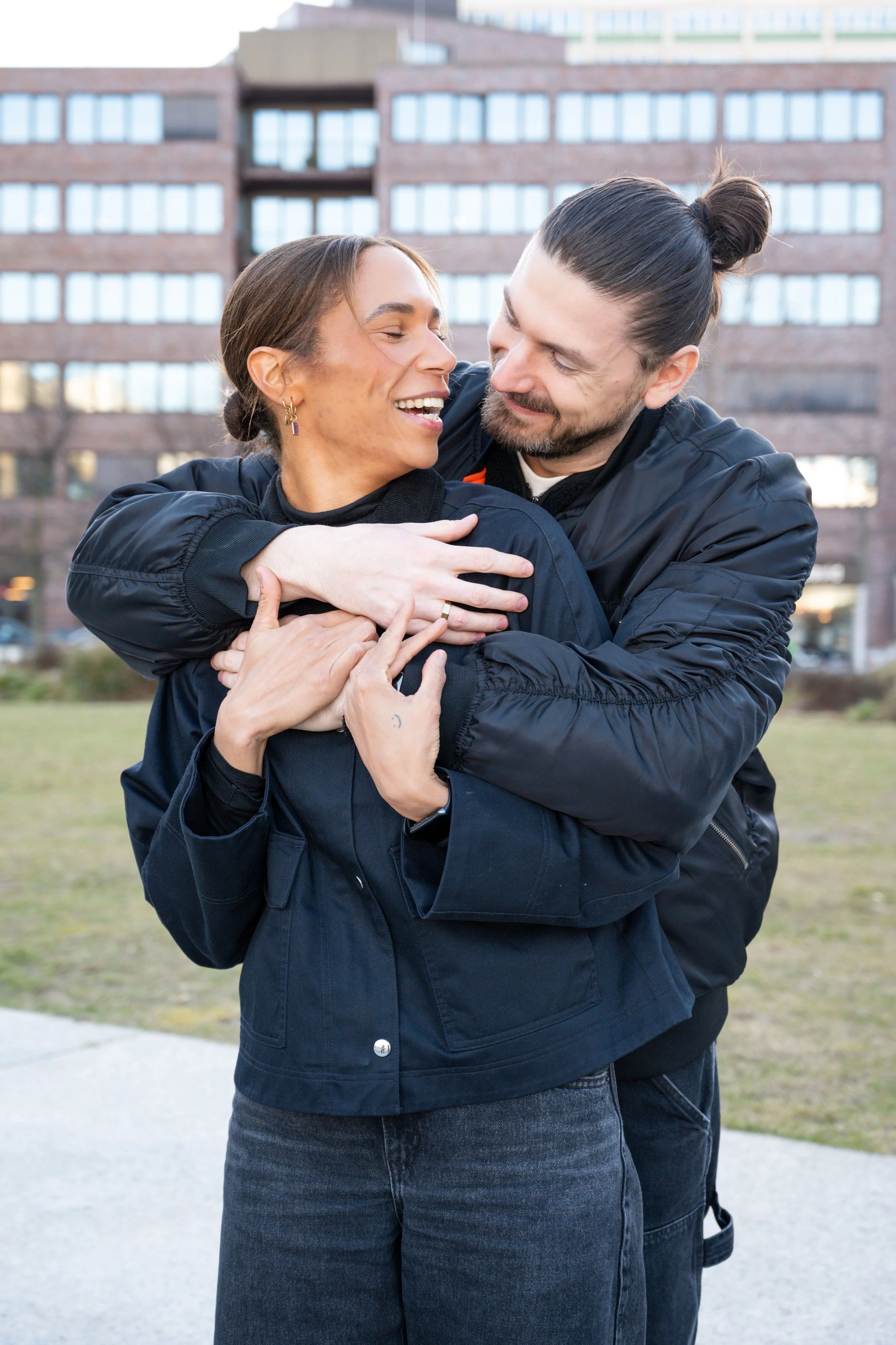 A smiling couple embracing outdoors, with a woman wearing a black jacket and a man in a black bomber jacket, in front of a modern brick building.