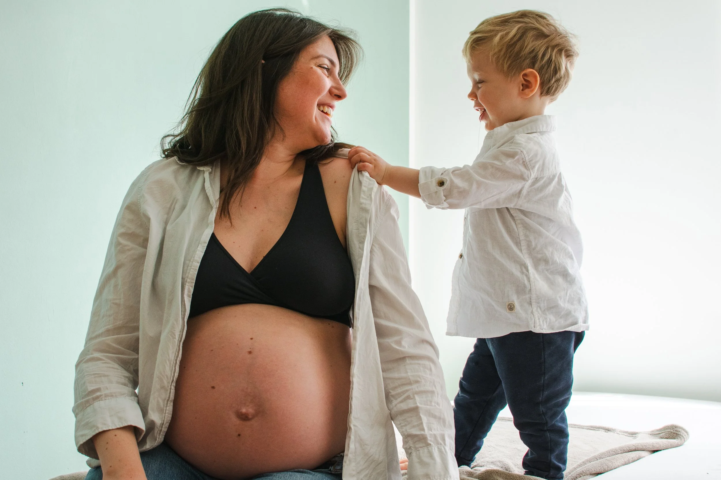 A pregnant woman smiling as she interacts with a young boy who is touching her shoulder, in a well-lit room.