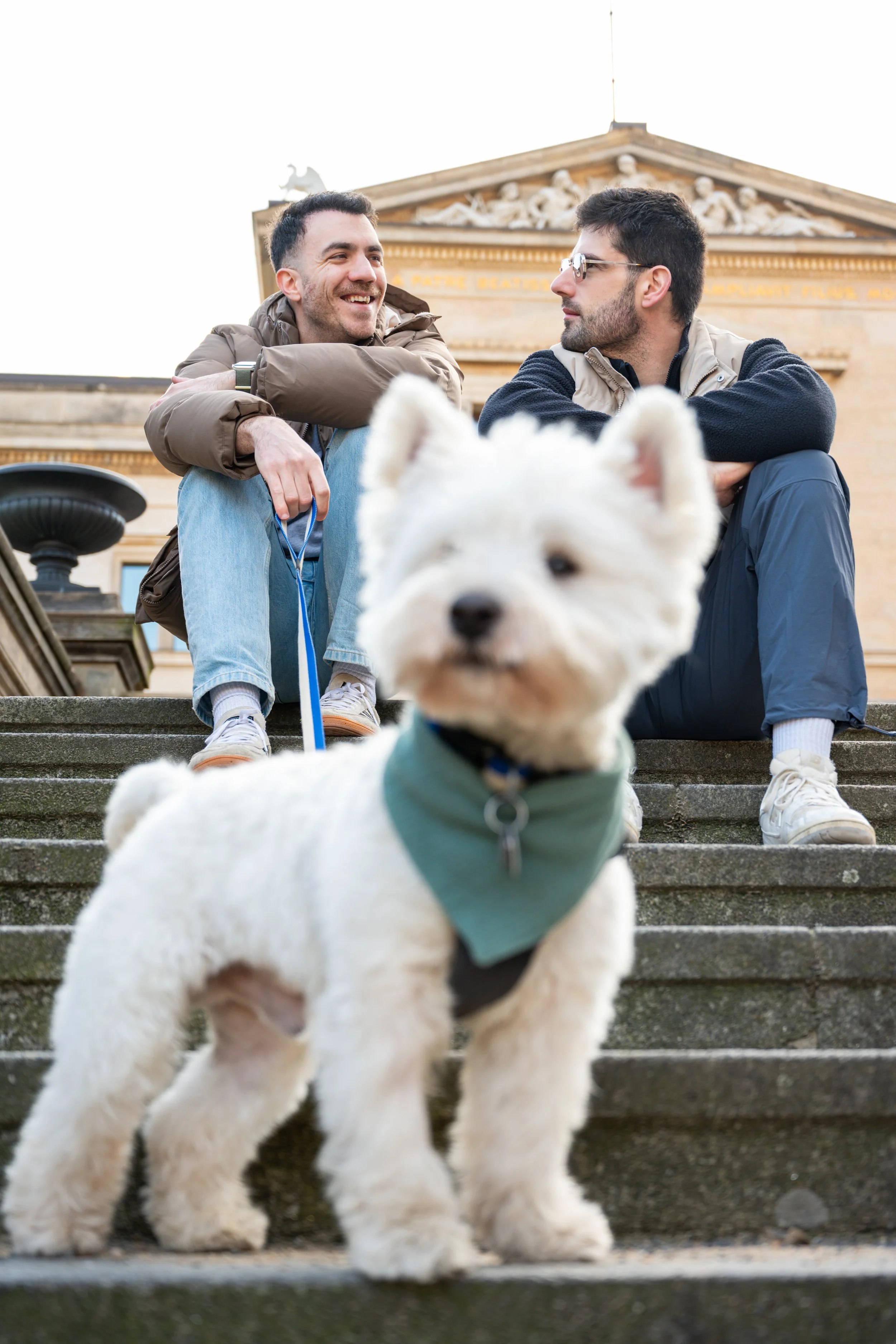 Two men sitting on stairs and talking, with a white dog in the foreground.