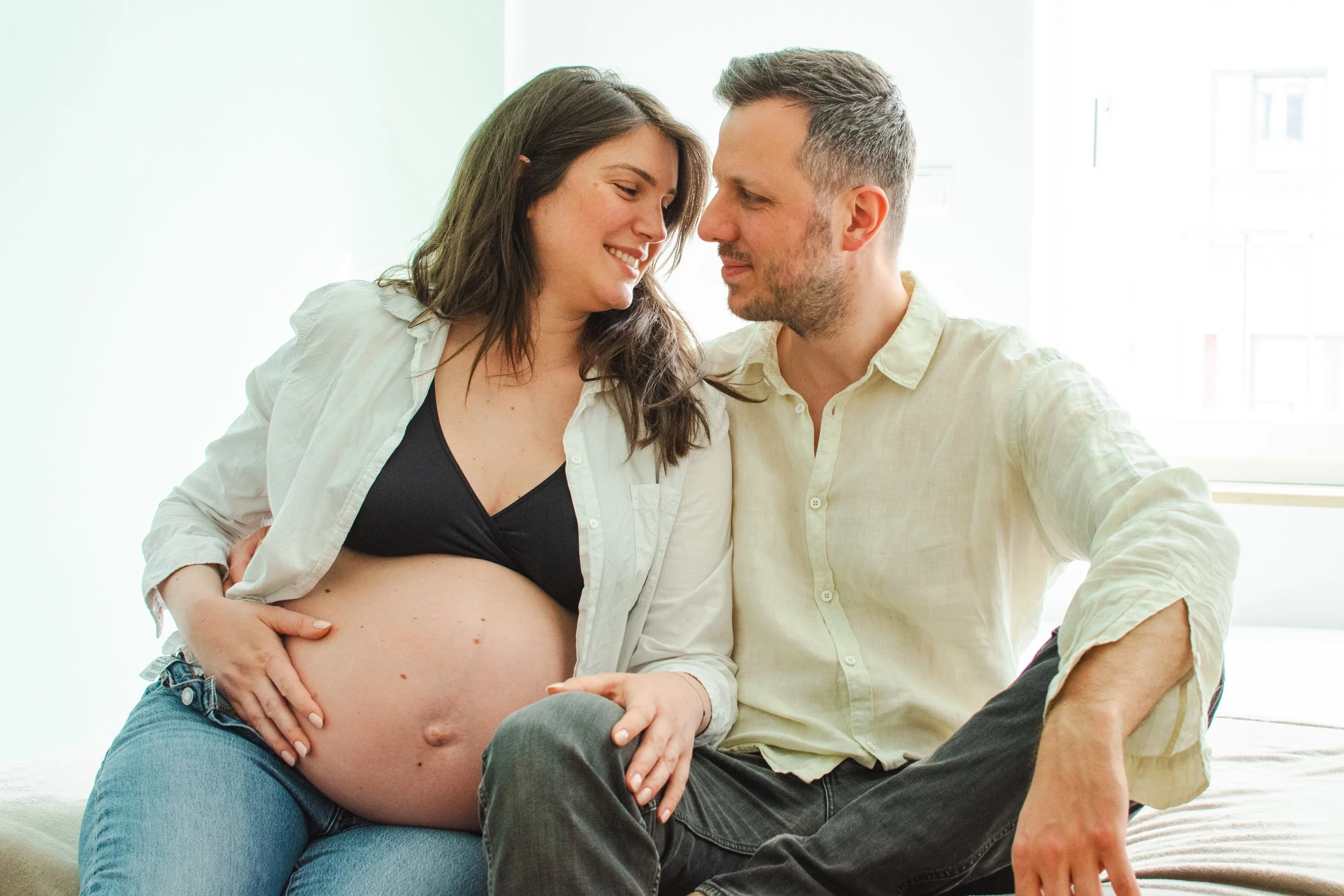 Pregnant woman and man sitting close together indoors, smiling at each other.