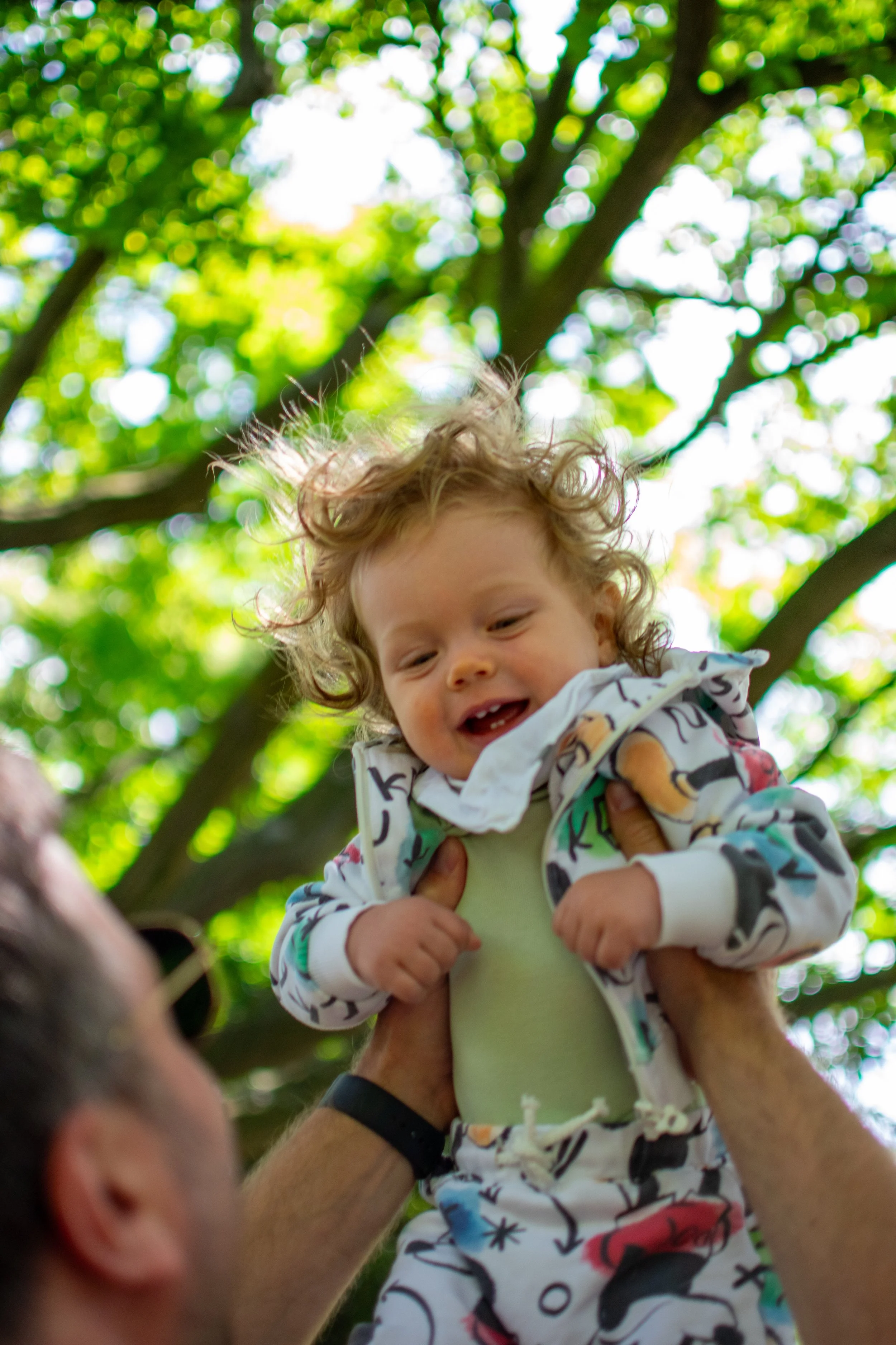 A young child with curly hair being lifted outdoors by an adult, with green tree leaves and sunlight in the background.