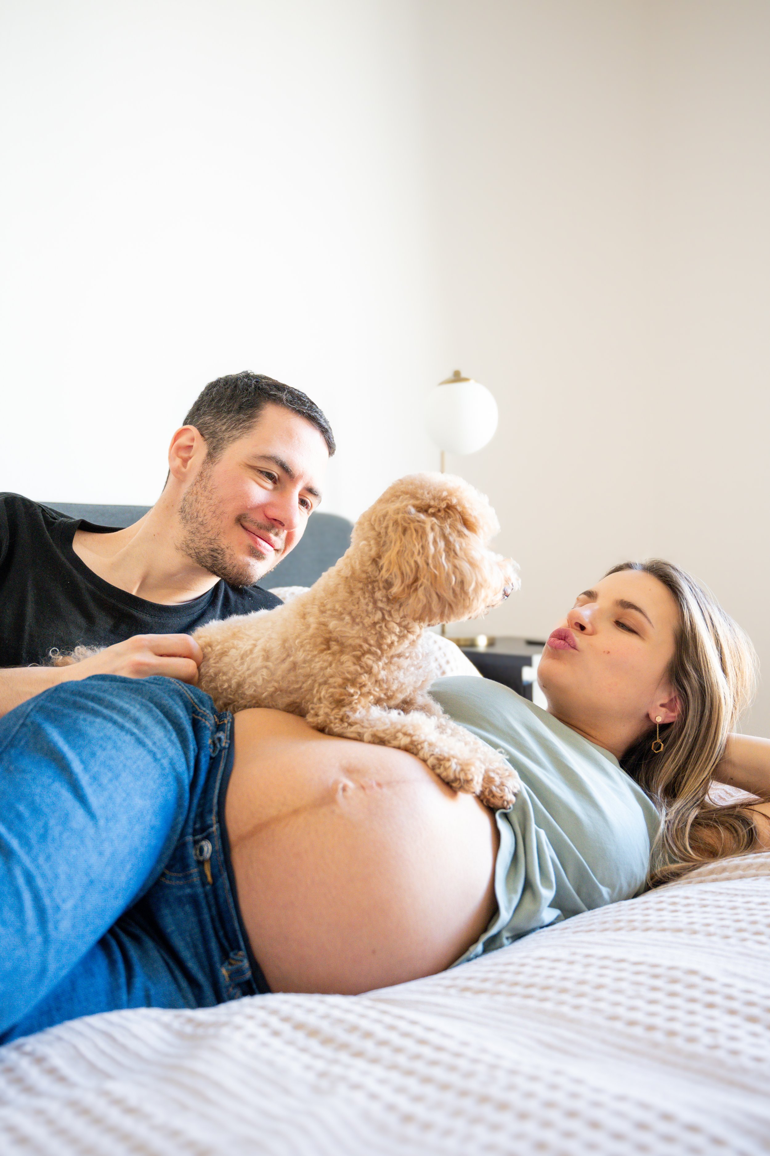 A expecting woman lying on a bed with a man and a small poodle dog, in a bright, cozy room.