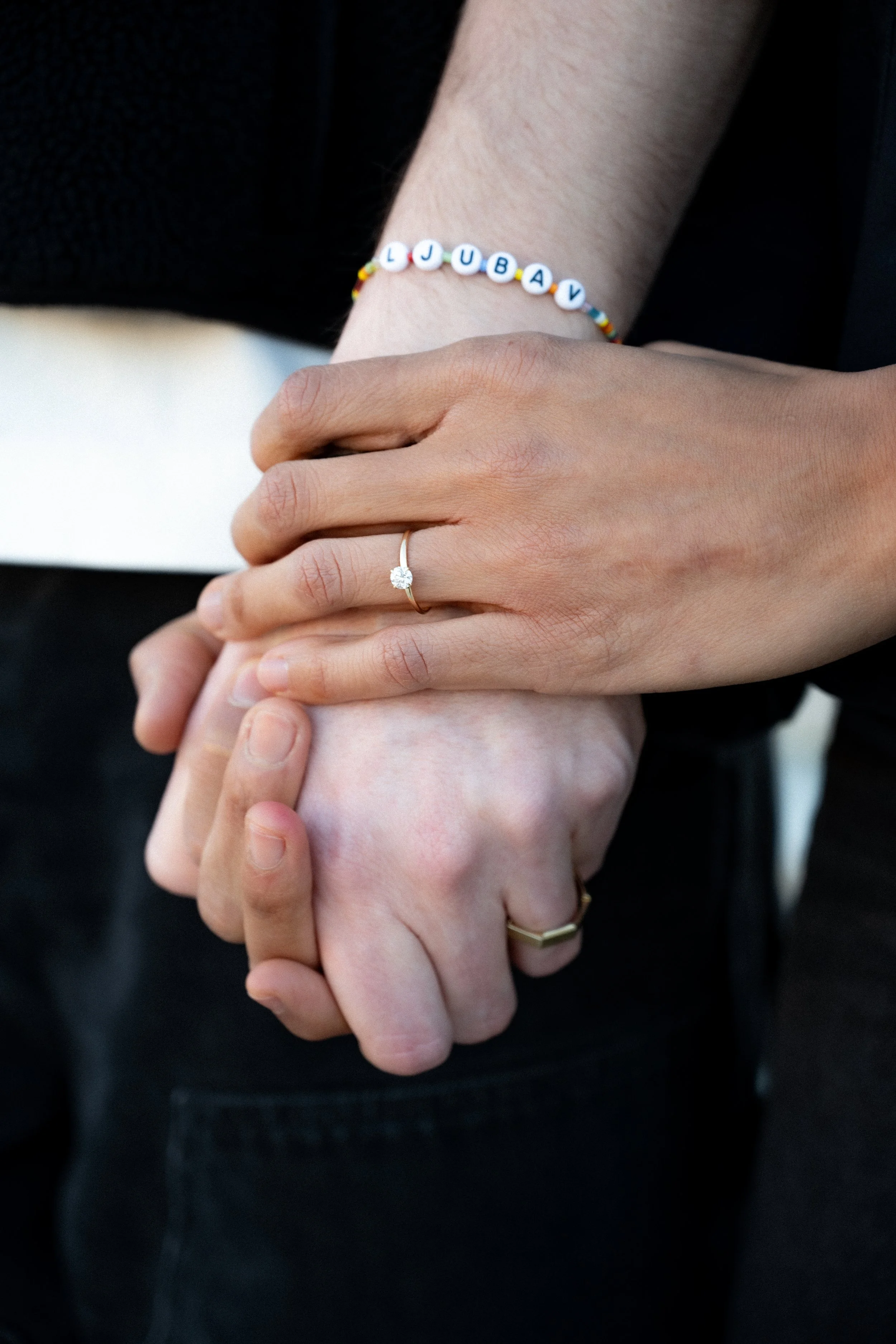 Close-up of two people holding hands, one wearing a beaded bracelet spelling out 'LJUBAV' and a diamond engagement ring, the other wearing a gold ring, dark clothing in the background.