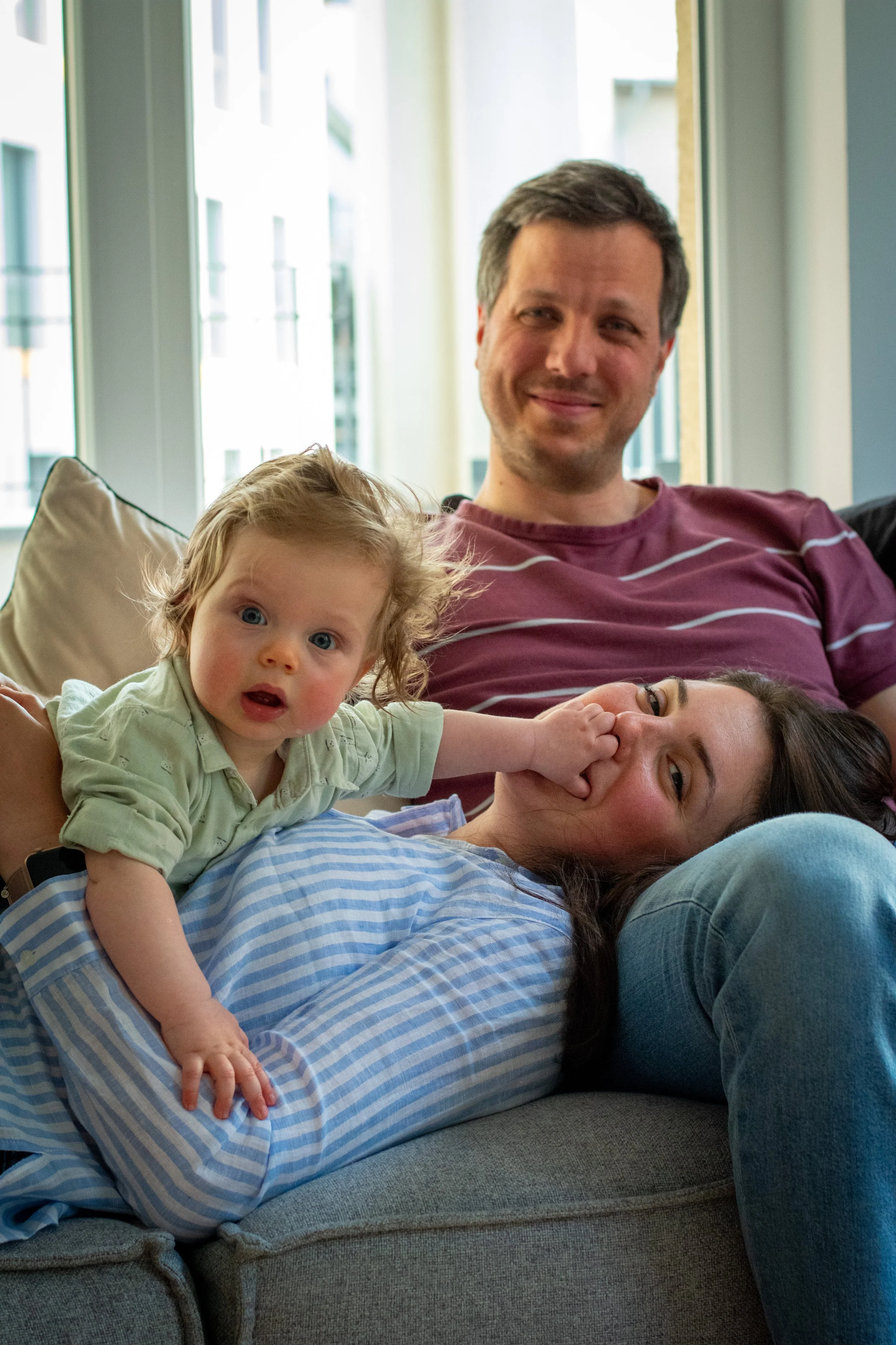 A family sitting on a couch near a window, with a young girl touching her mother's nose and a man smiling in the background.