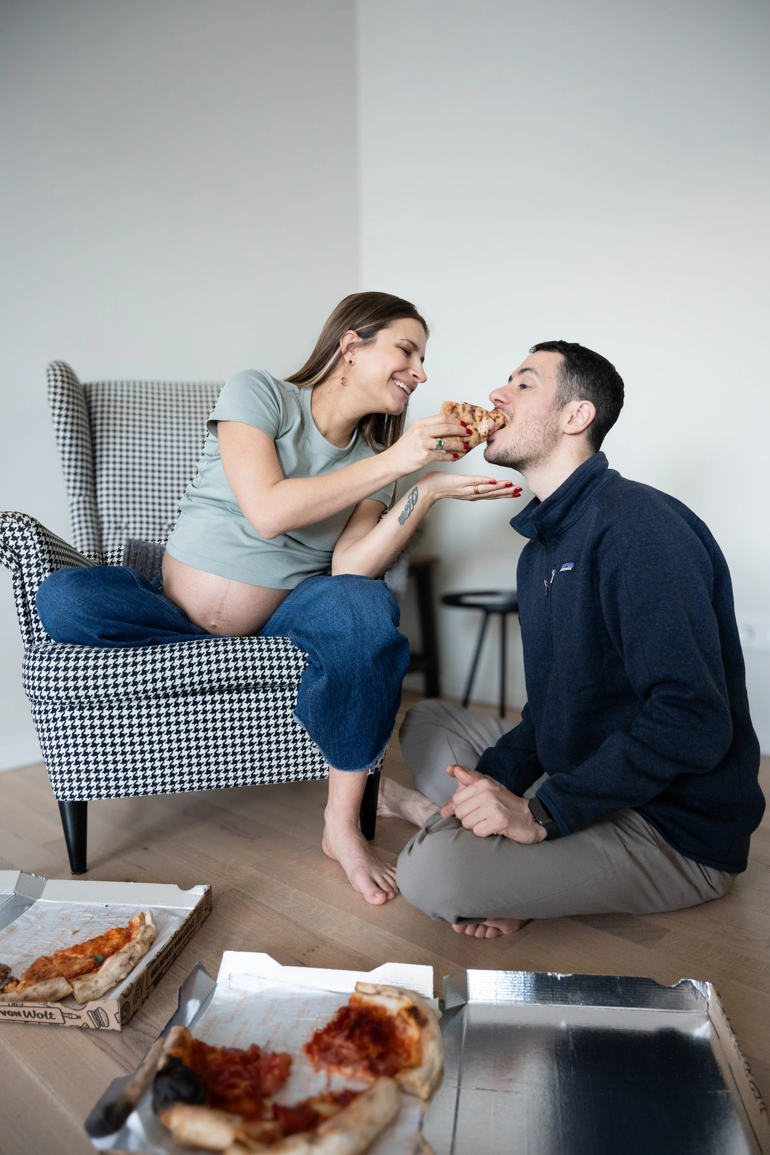 A woman sitting on a checkered armchair feeding pizza to a man kneeling on the floor, with pizza boxes on the floor.