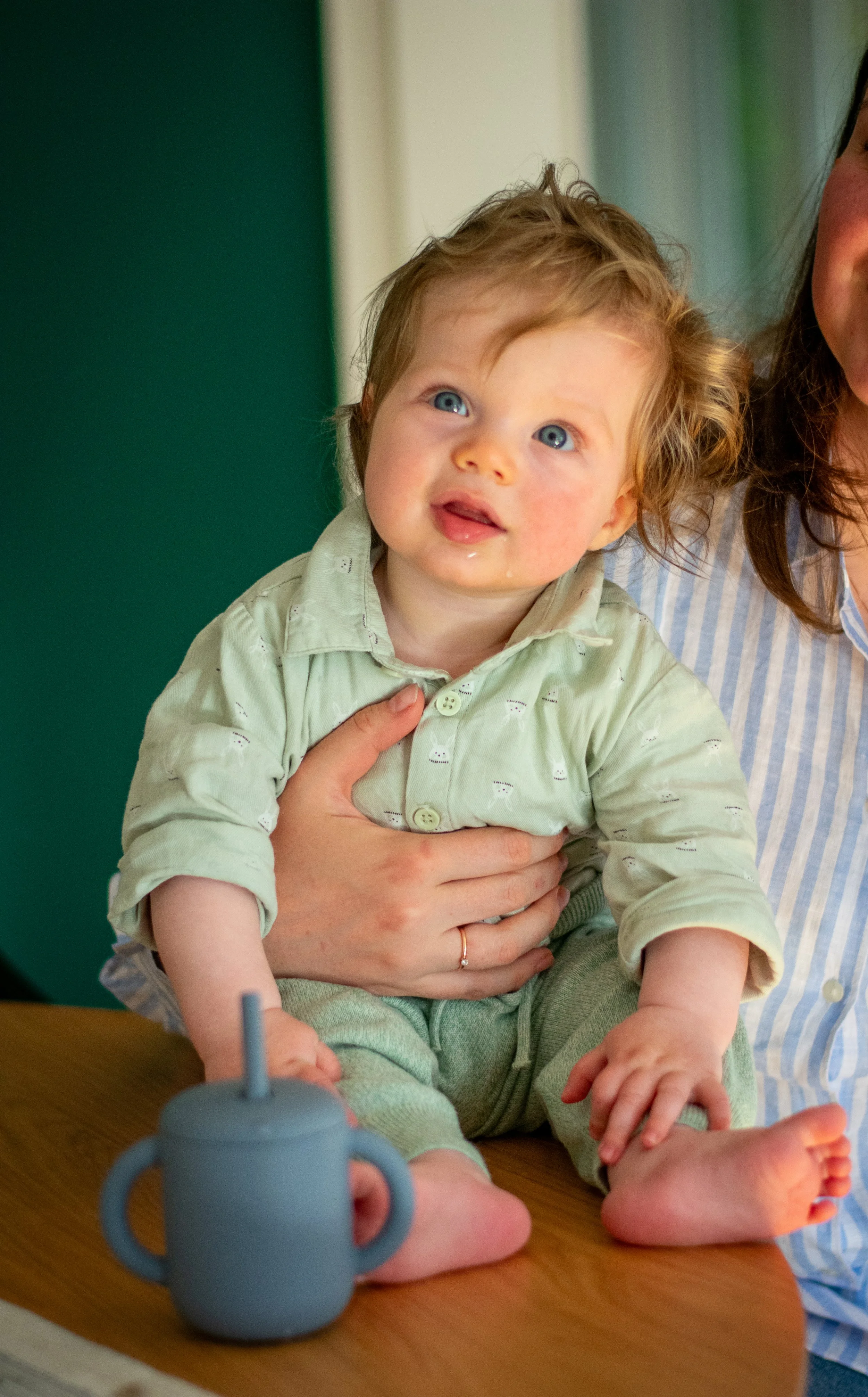 A young child with curly blonde hair and blue eyes sitting on a table, being held by a woman. The child is wearing a light green shirt and pants, and has a slight smile. There is a blue sippy cup on the table in front of them.