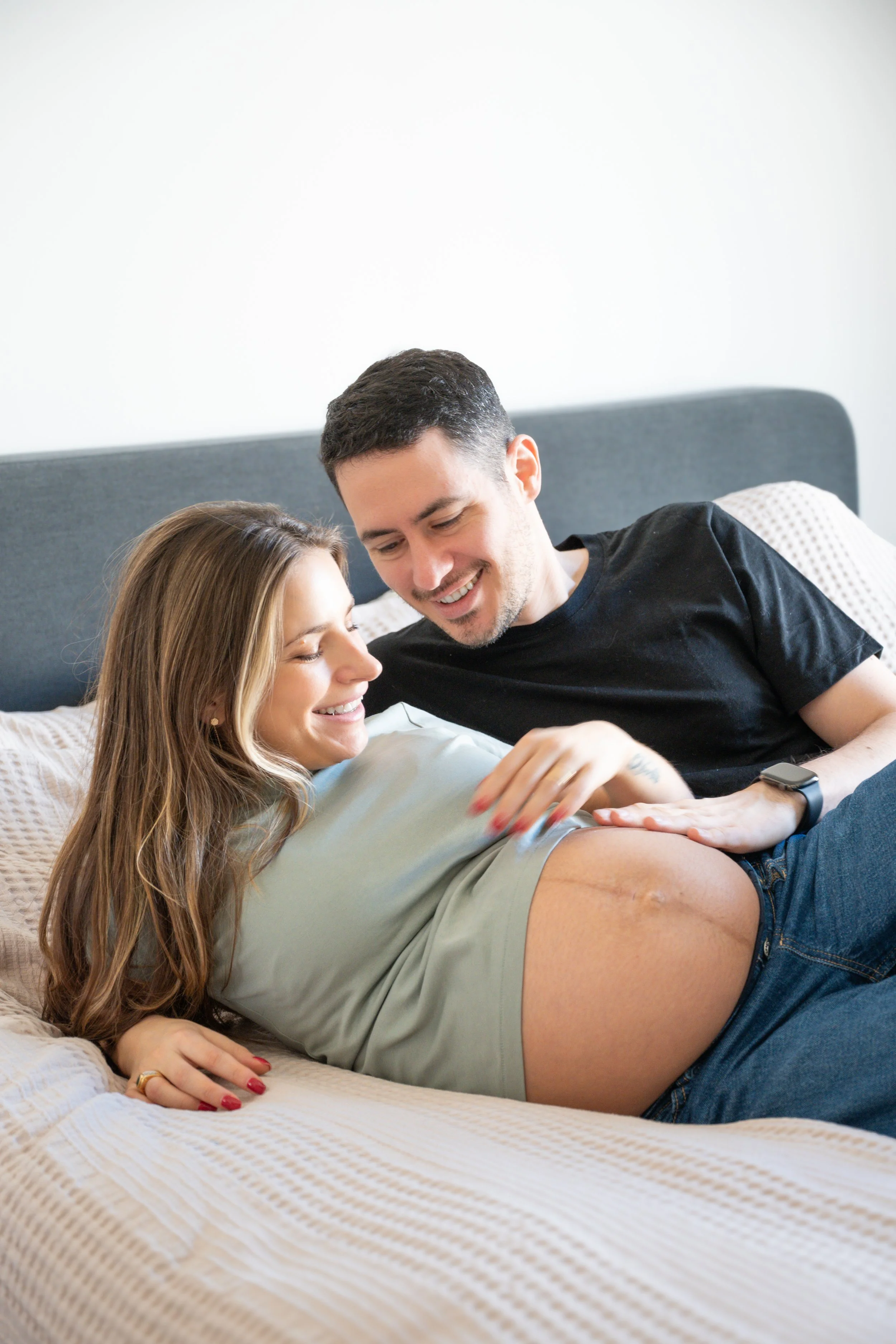 A happy couple in bed with a pregnant woman smiling and touching her belly, a man leaning over her with a smile.