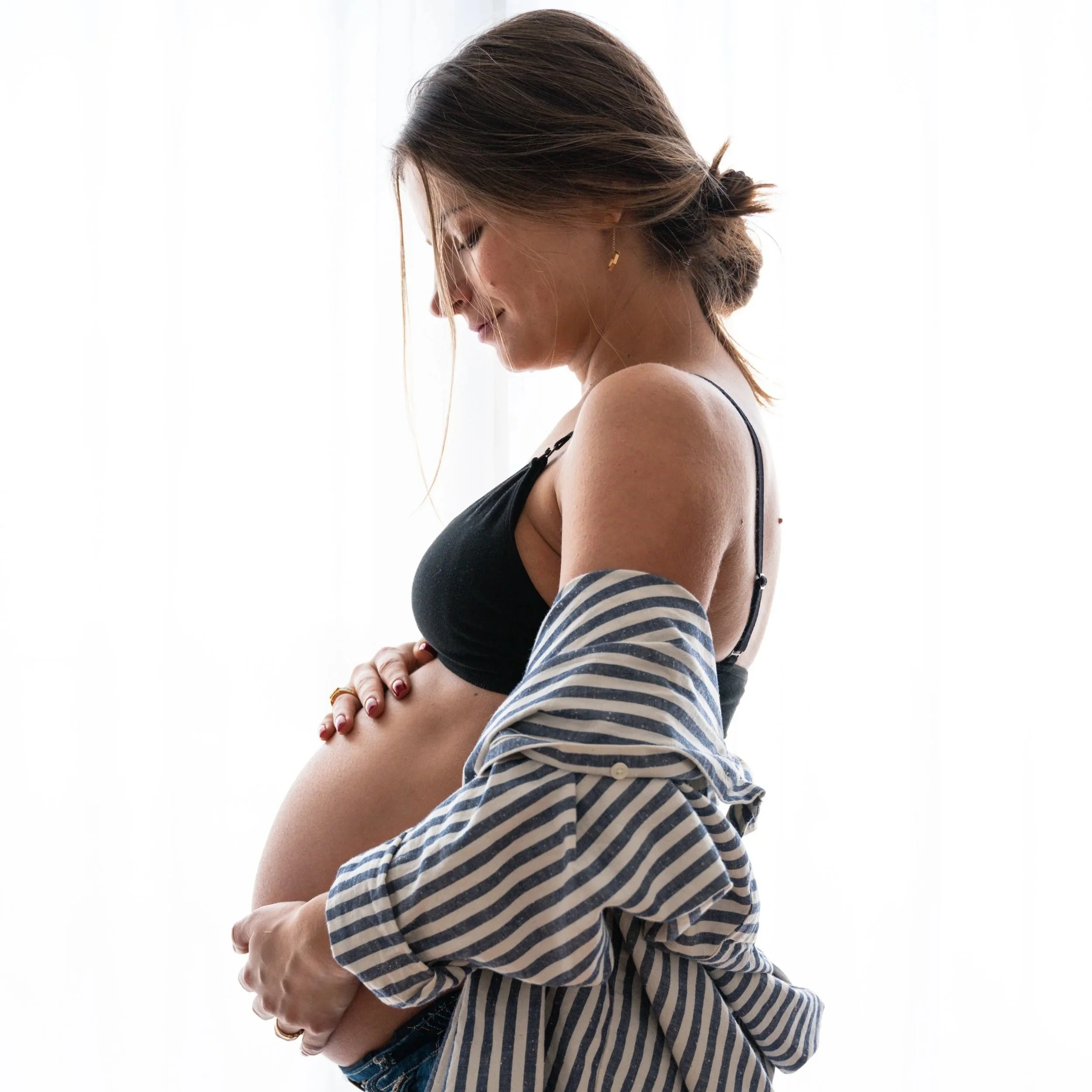 Pregnant woman smiling, wearing a black bra, with a striped shirt draped over her arm, standing near a window with white curtains.