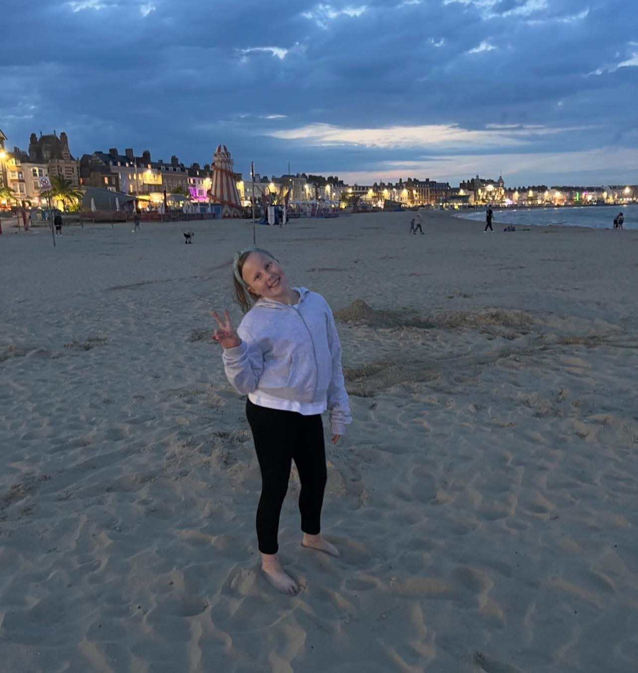 A smiling girl in a white hoodie and black pants making a peace sign on a sandy beach at dusk. In the background, there are illuminated buildings, a carousel, and a cloudy sky over the shoreline.