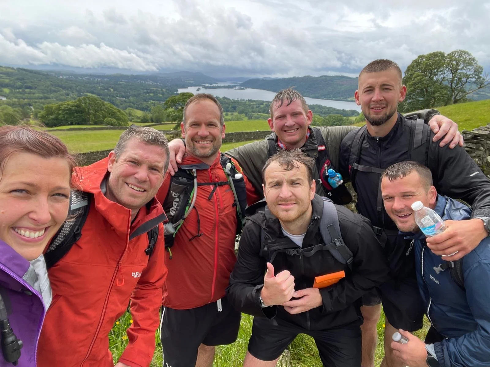 Group of eight hikers posing together outdoors with a scenic lake and green hills in the background, cloudy sky overhead.