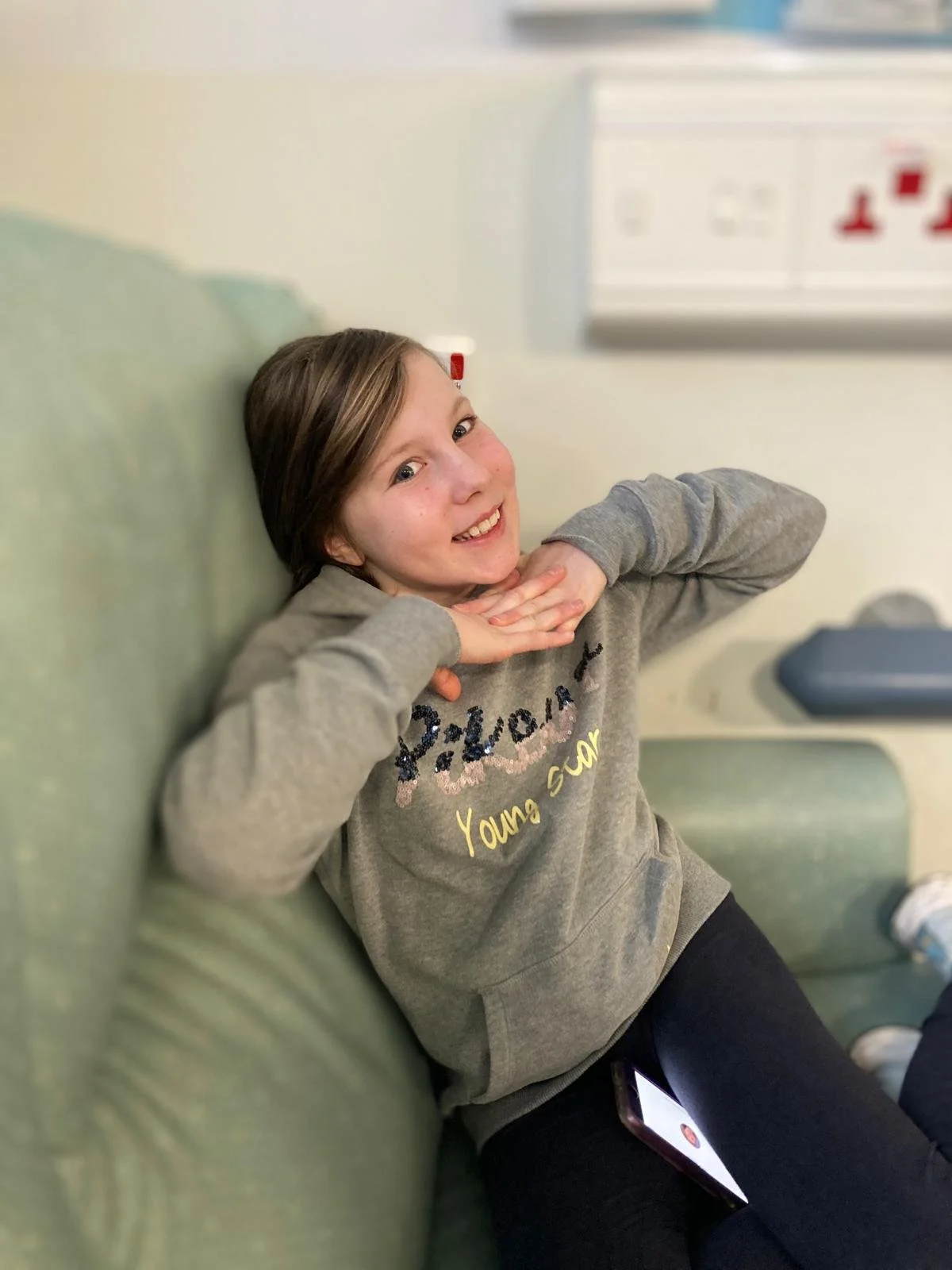 A girl with brown hair lying on a green couch, smiling at the camera, wearing a gray hoodie with text, and resting her hands under her chin. There is a smartphone on her lap.