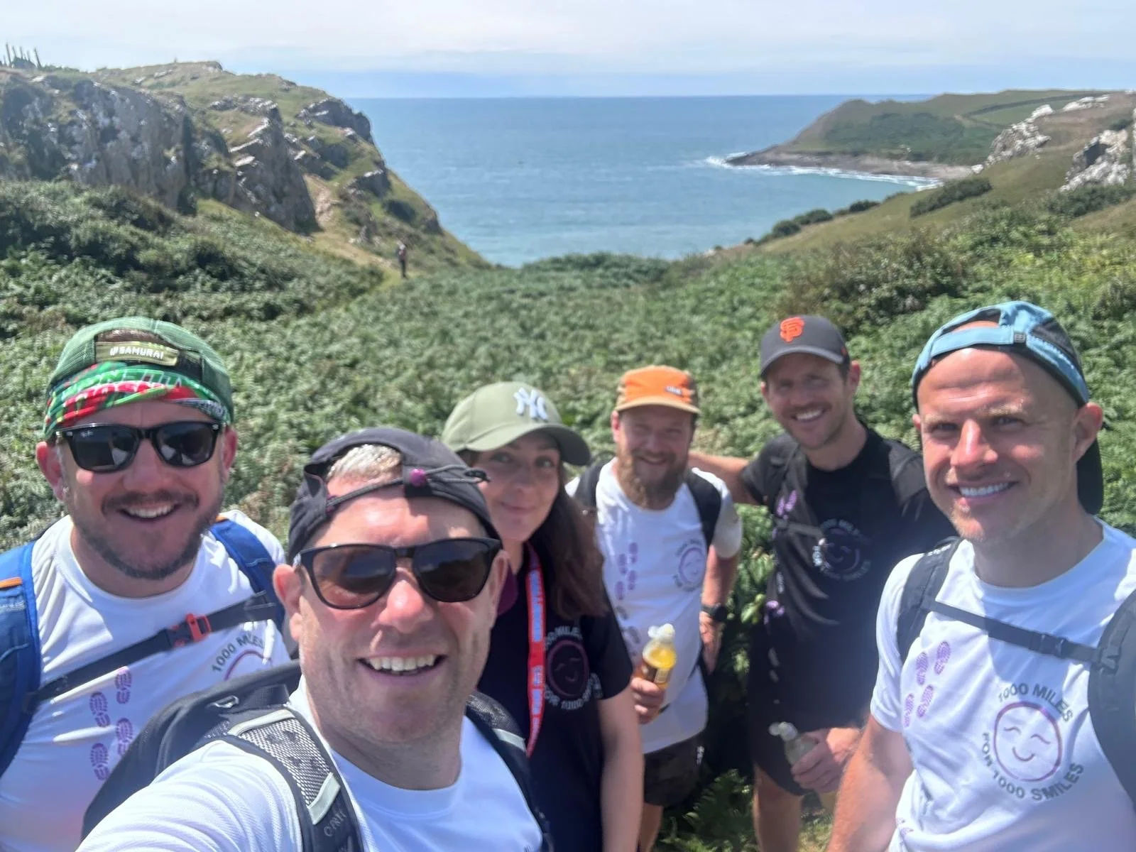 Group of six people hiking outdoors on a trail near the coast with cliffs and ocean in the background.