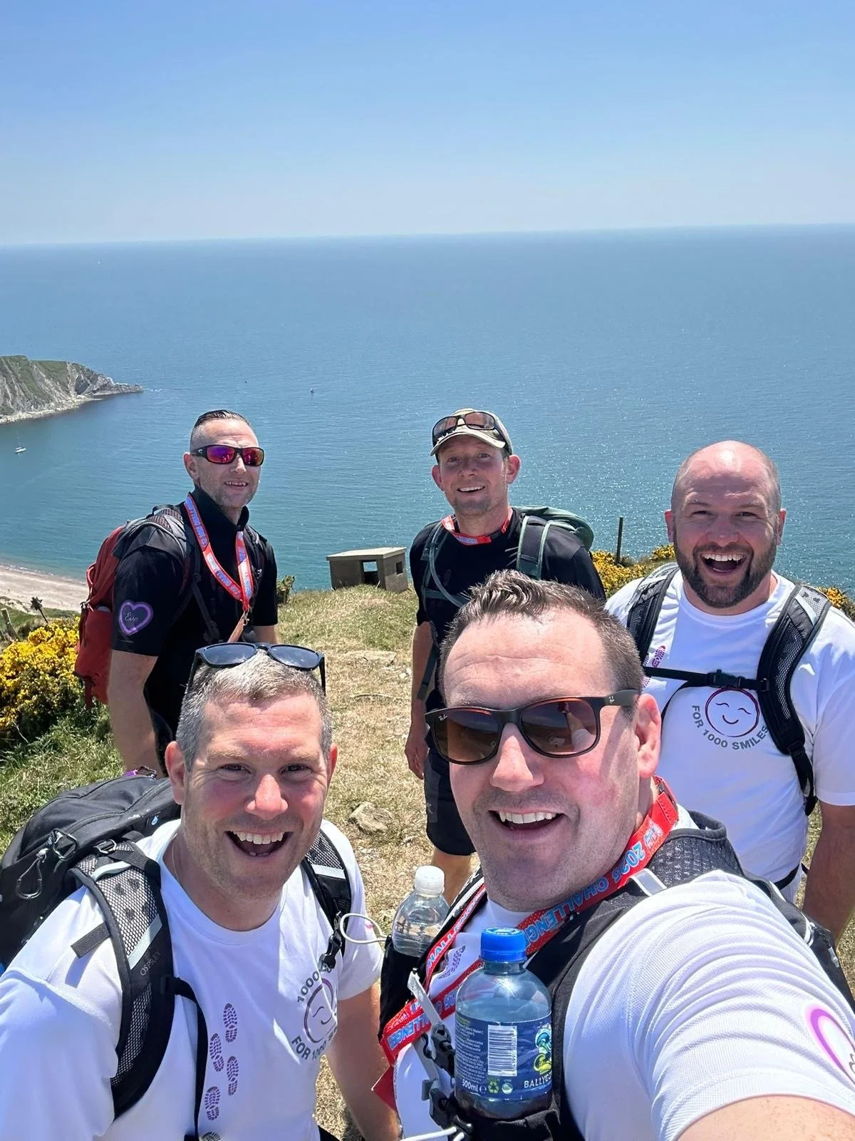 A group of six men taking a selfie on a coastal cliff during a sunny day, with ocean and sky in the background, dressed in hiking gear and smiling.