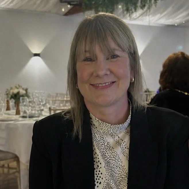 A woman with shoulder-length blonde hair smiling at a formal event, seated at a decorated table with white tablecloths and floral centerpieces.