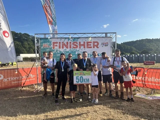 Group of people, including children, standing under a banner that says 'FINISHER' after completing a 50 kilometer race, outdoors with a clear sky and flags.