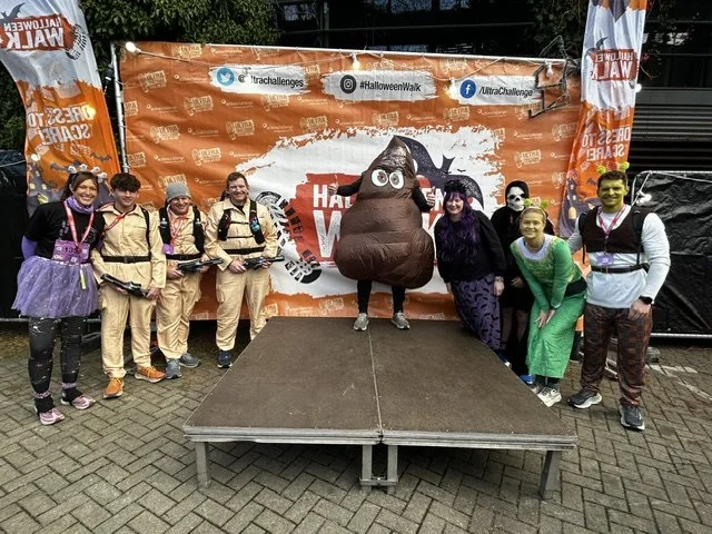 People in Halloween costumes, including a pooping emoji mascot, standing on a stage in front of a Halloween event backdrop with orange and black decorations.