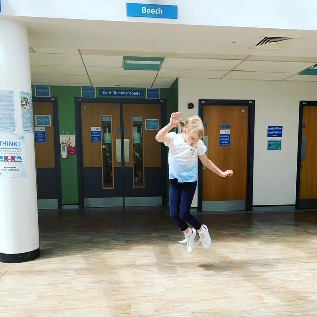 Child jumping with arms up in a hallway at a healthcare facility, with doors and signs indicating a treatment suite in the background.