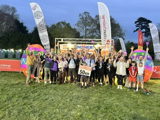 Group of people celebrating at the finish line of a race, holding a '50 KM' sign, with colorful inflatable decorations and banners overhead in an outdoor park setting.