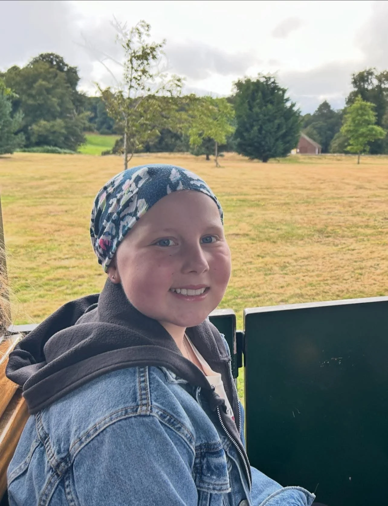 Smiling girl with a headscarf sitting outdoors with trees and a field in the background.