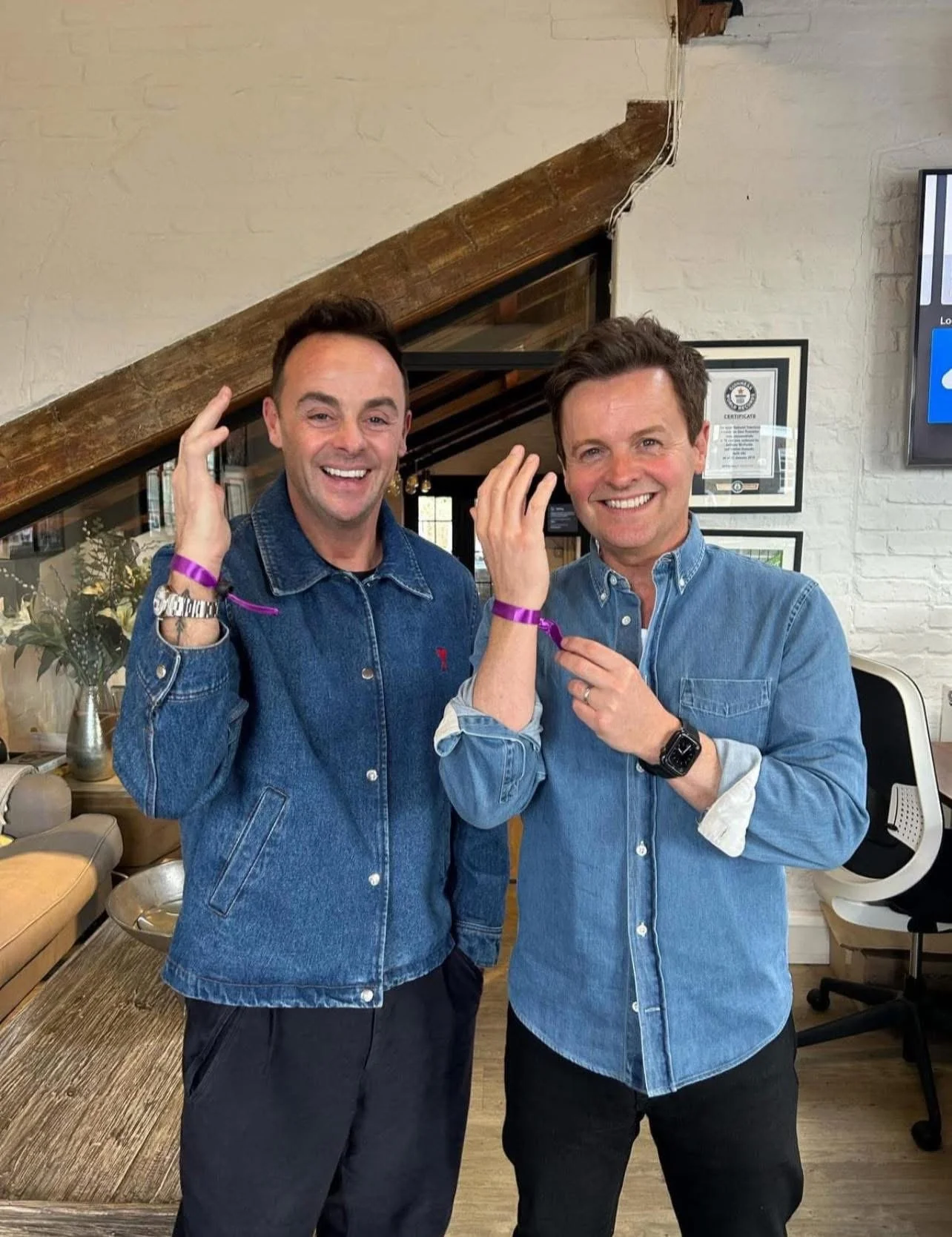 Two men wearing denim shirts smiling, standing indoors, each with a purple wristband and touching their ears.