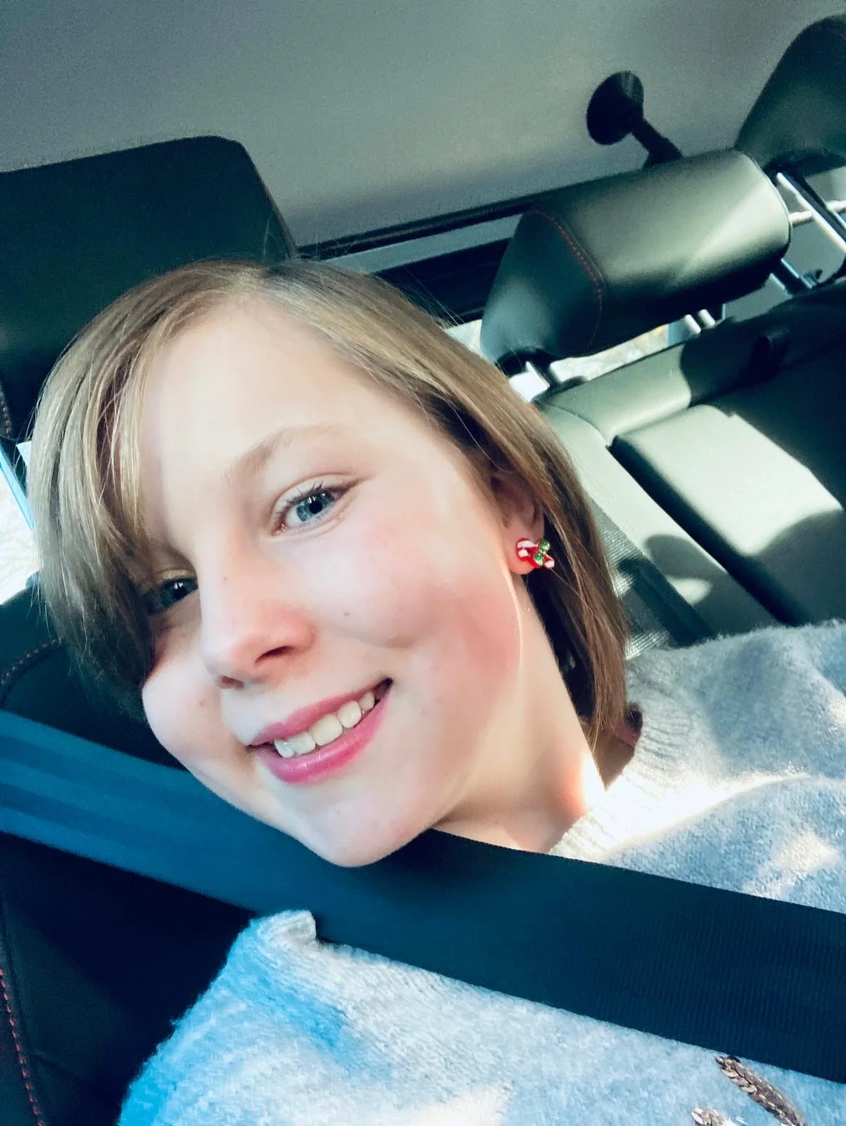 A young woman with short light brown hair and blue eyes smiling while sitting in a car, wearing Christmas-themed earrings.