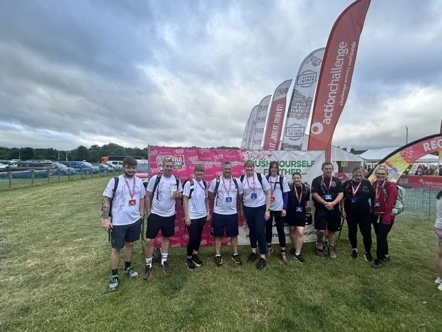Group of nine people standing outdoors in front of pink banners at a running event, all wearing athletic clothes and race bibs.